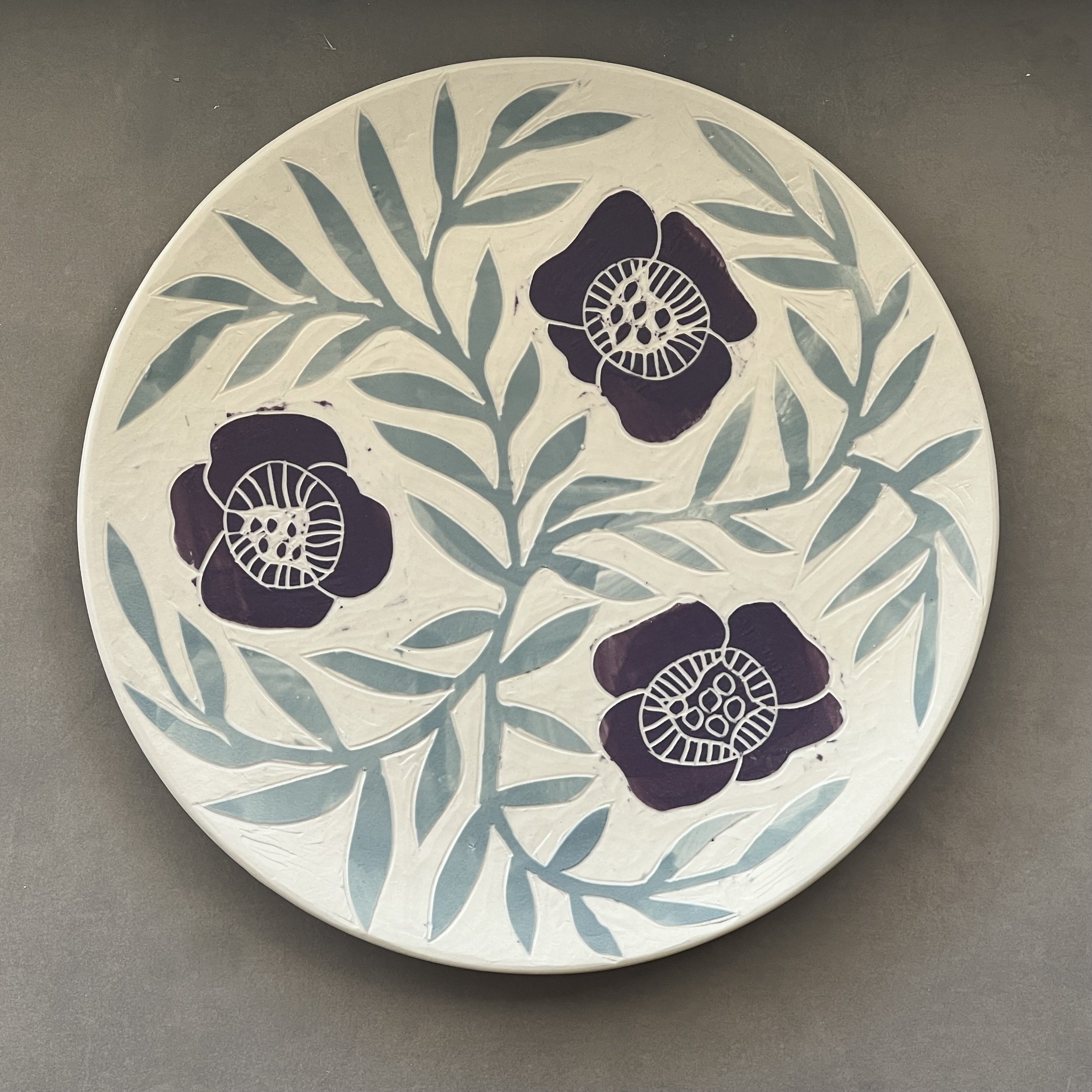 Dimity Kidston sgraffito hand carved ceramic plate with cut-out floral and leaf patterns, featuring black flowers with intricate white designs, on a gray surface.