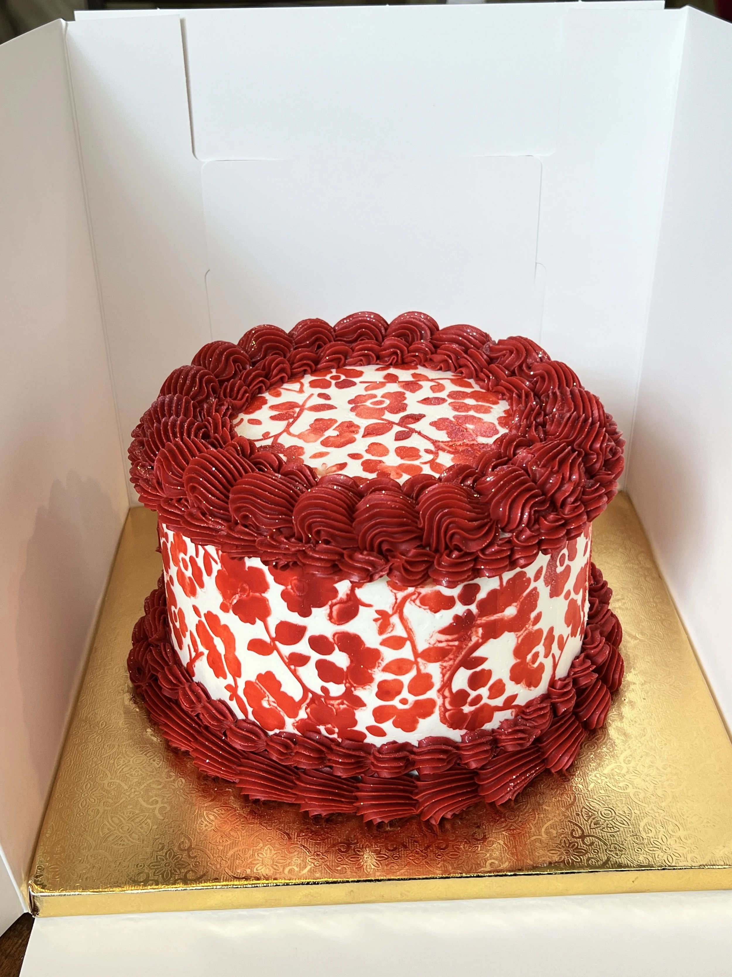 A round layered cake with white fondant decorated with red floral patterns and surrounded by red piped frosting design, placed on a gold-colored base inside a white display box.