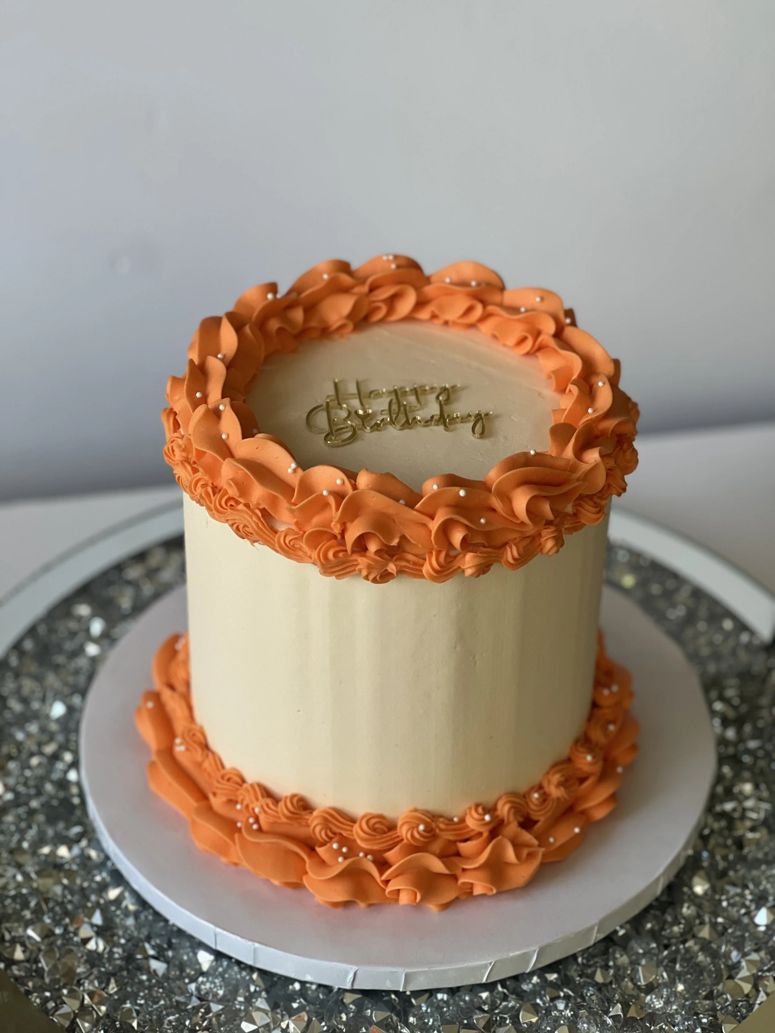 A birthday cake decorated with orange frosting and small white pearl-like sprinkles, with "Happy Birthday" written on top in gold.