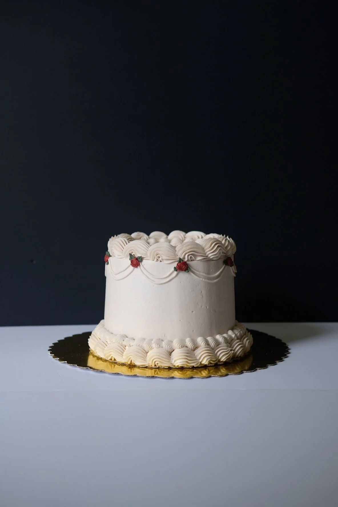 A white, round celebration cake with decorative icing patterns, red icing roses, and green leaves, placed on a gold cake board against a black background.