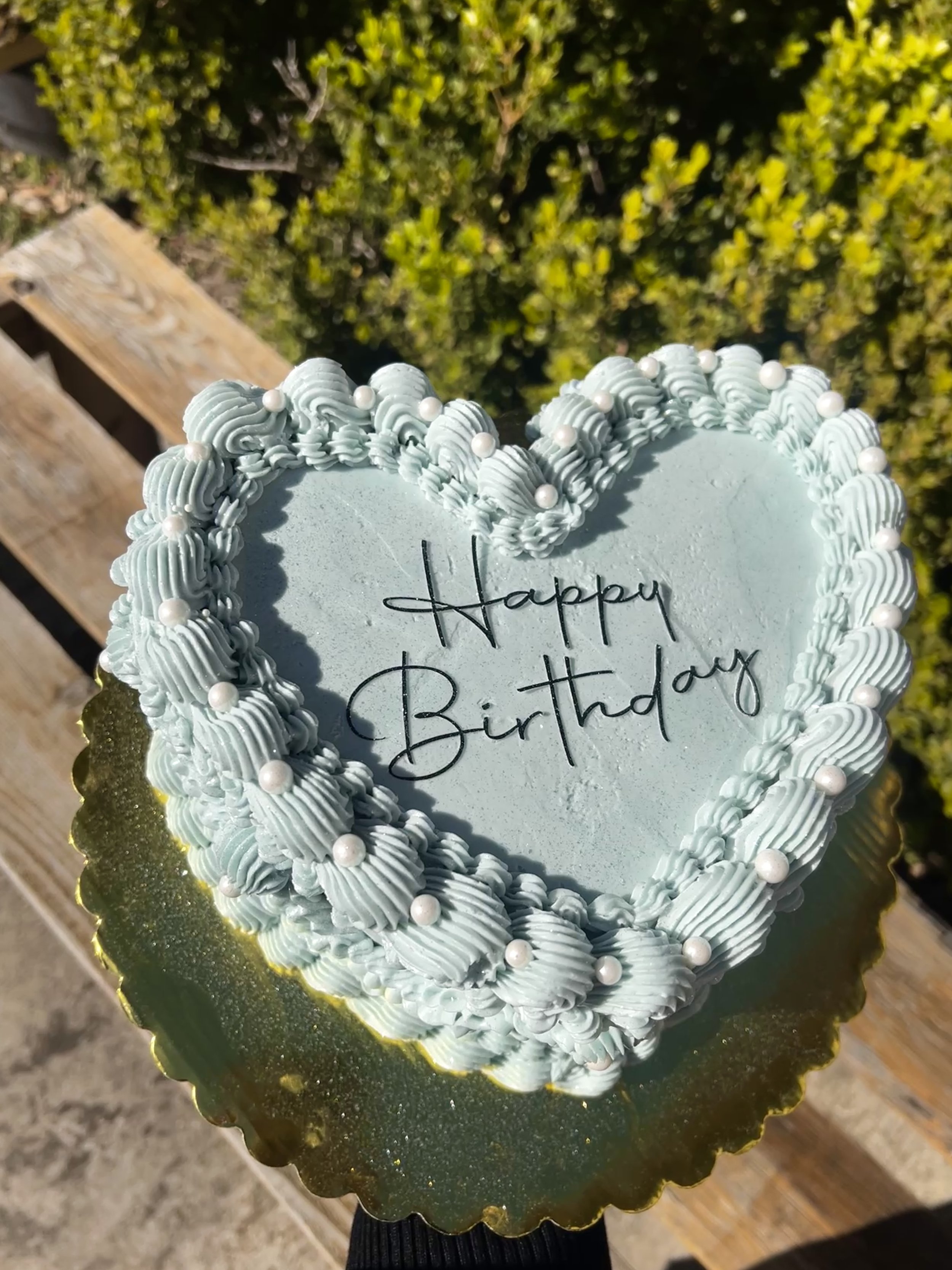 A heart-shaped birthday cake decorated with light blue icing, white pearls, and swirled piping, with a message written in black icing that says "Happy Birthday."