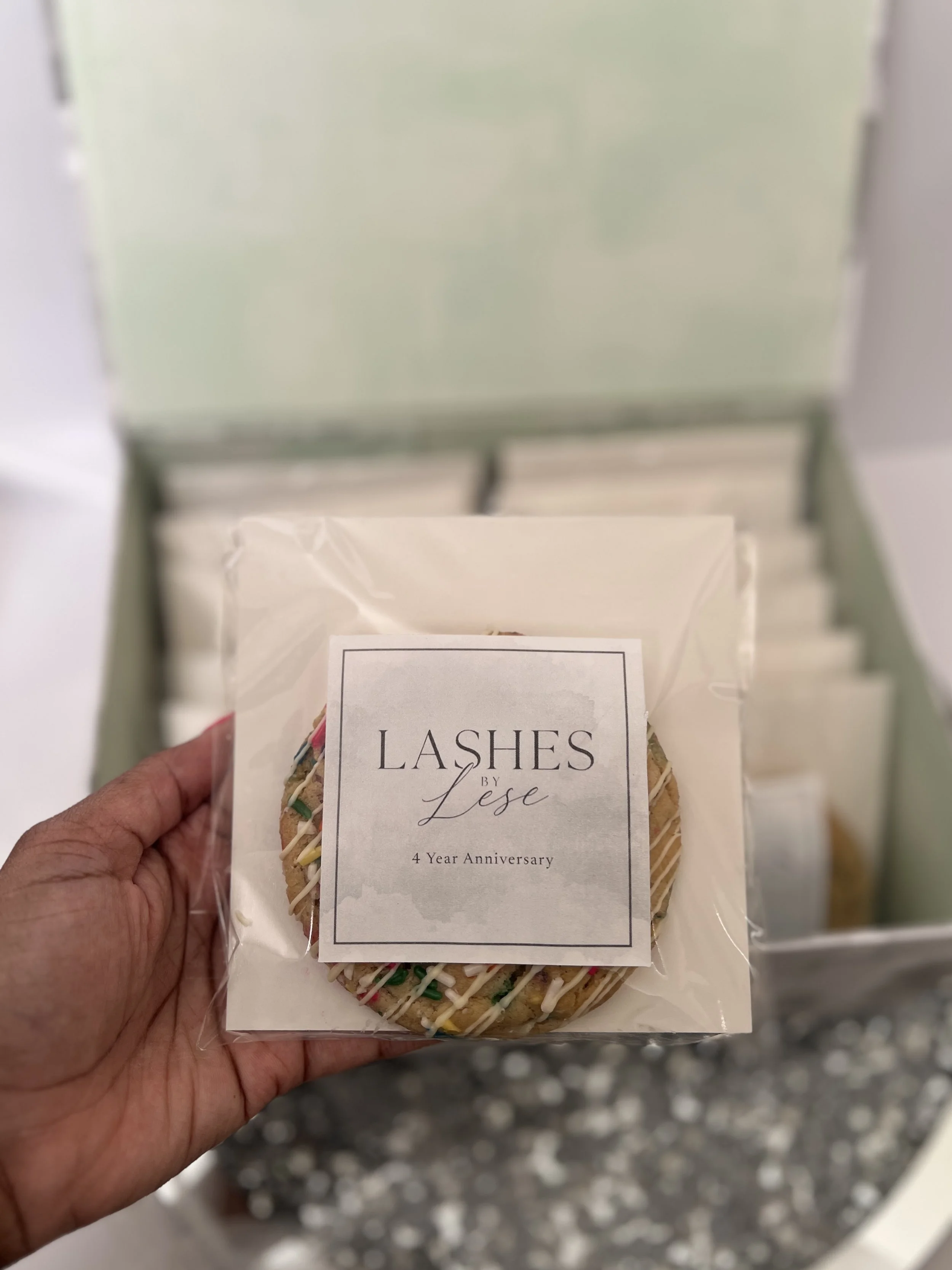 Close-up of a hand holding a packaged cookie decorated with colorful sprinkles and white icing, with a label that says "Lashes by Lese 4 Year Anniversary."