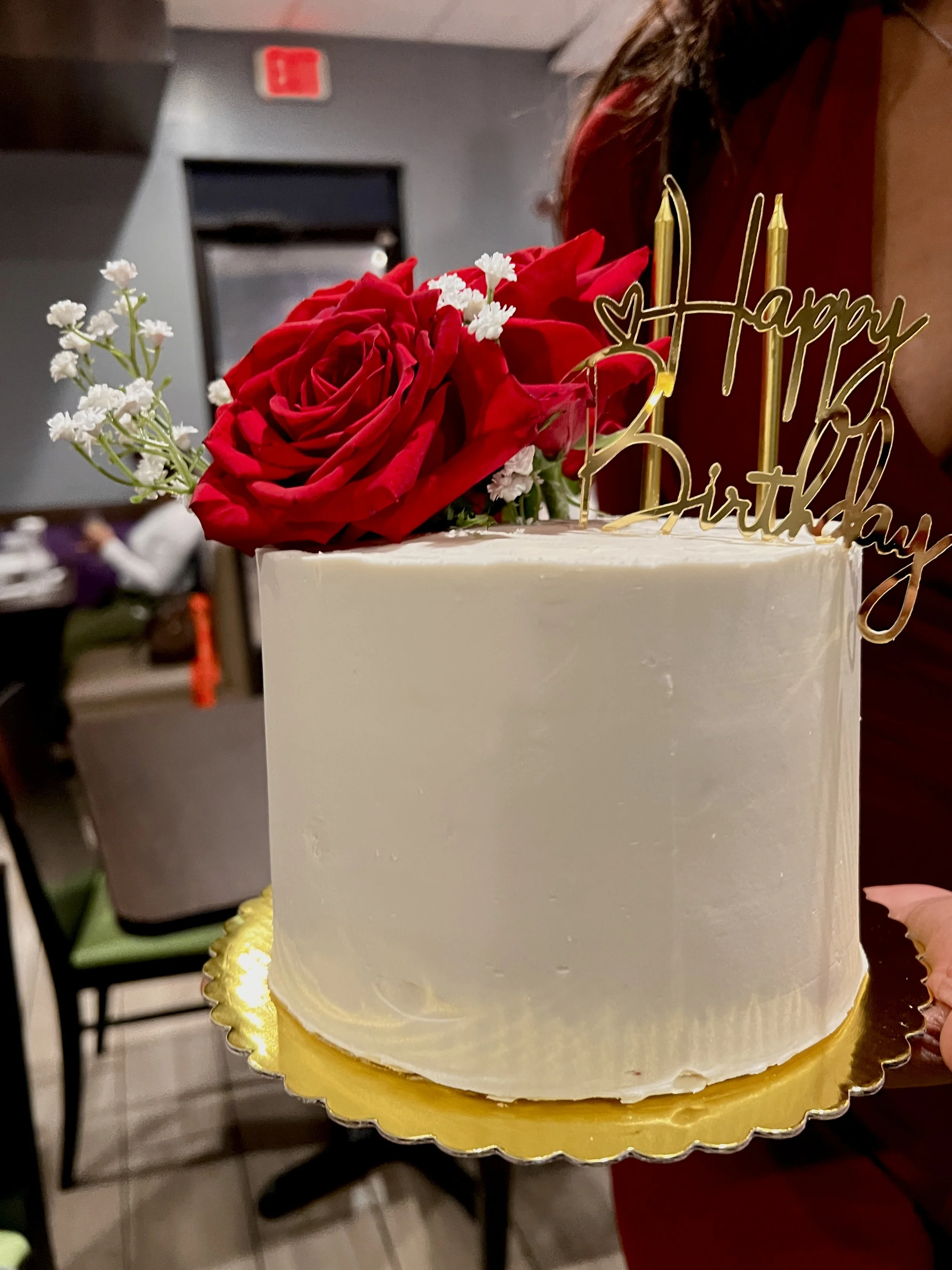 A birthday cake decorated with white frosting, topped with red roses and small white flowers, and a gold 'Happy Birthday' cake topper with candles.
