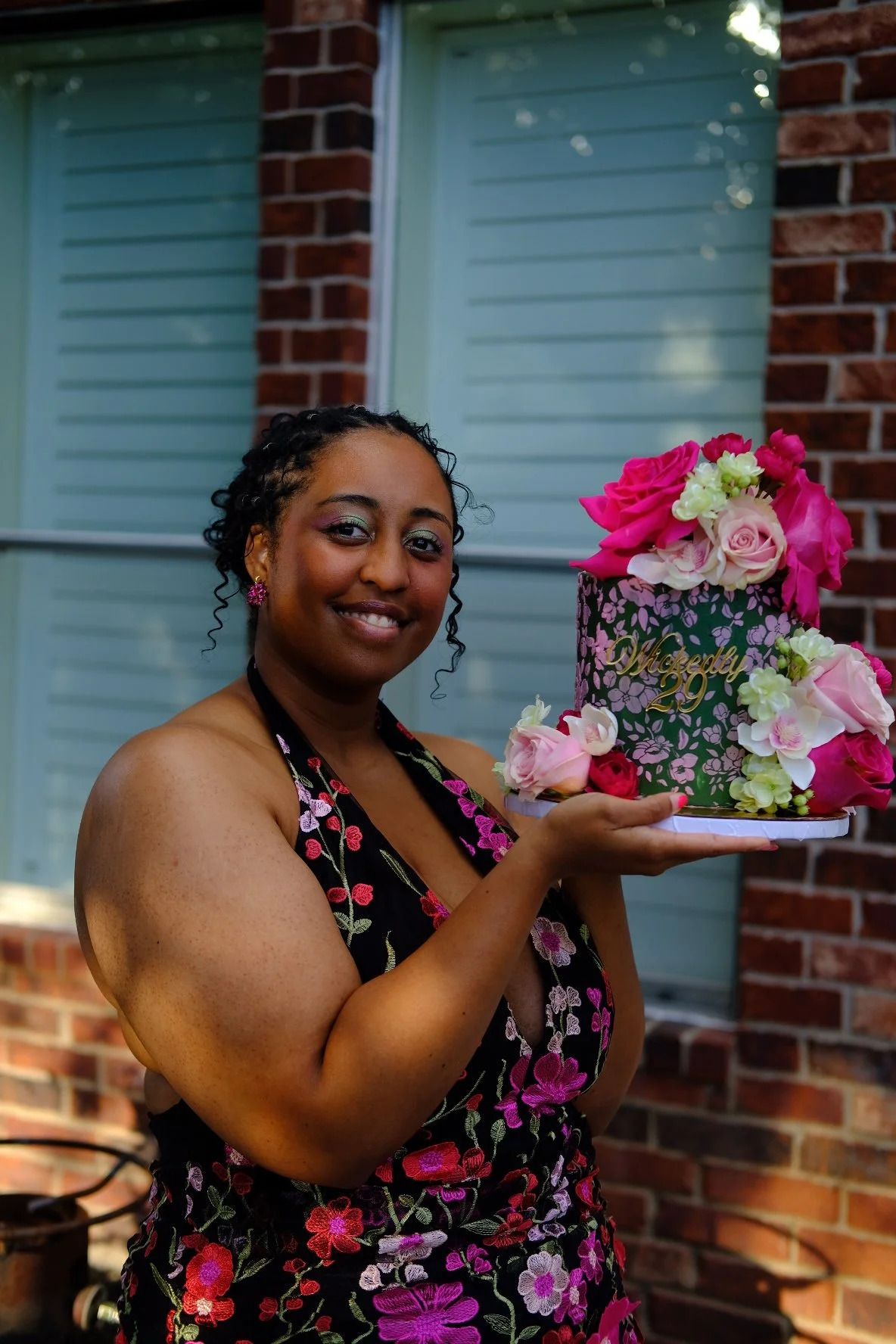 A woman with dark, curly hair and a floral dress smiles while holding a decorative cake adorned with pink and white flowers, with a brick house and green window shutters in the background.