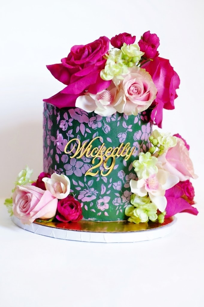 A floral cake decorated with pink and white roses, greenery, and the words 'Wickedly 29' in gold on the front.