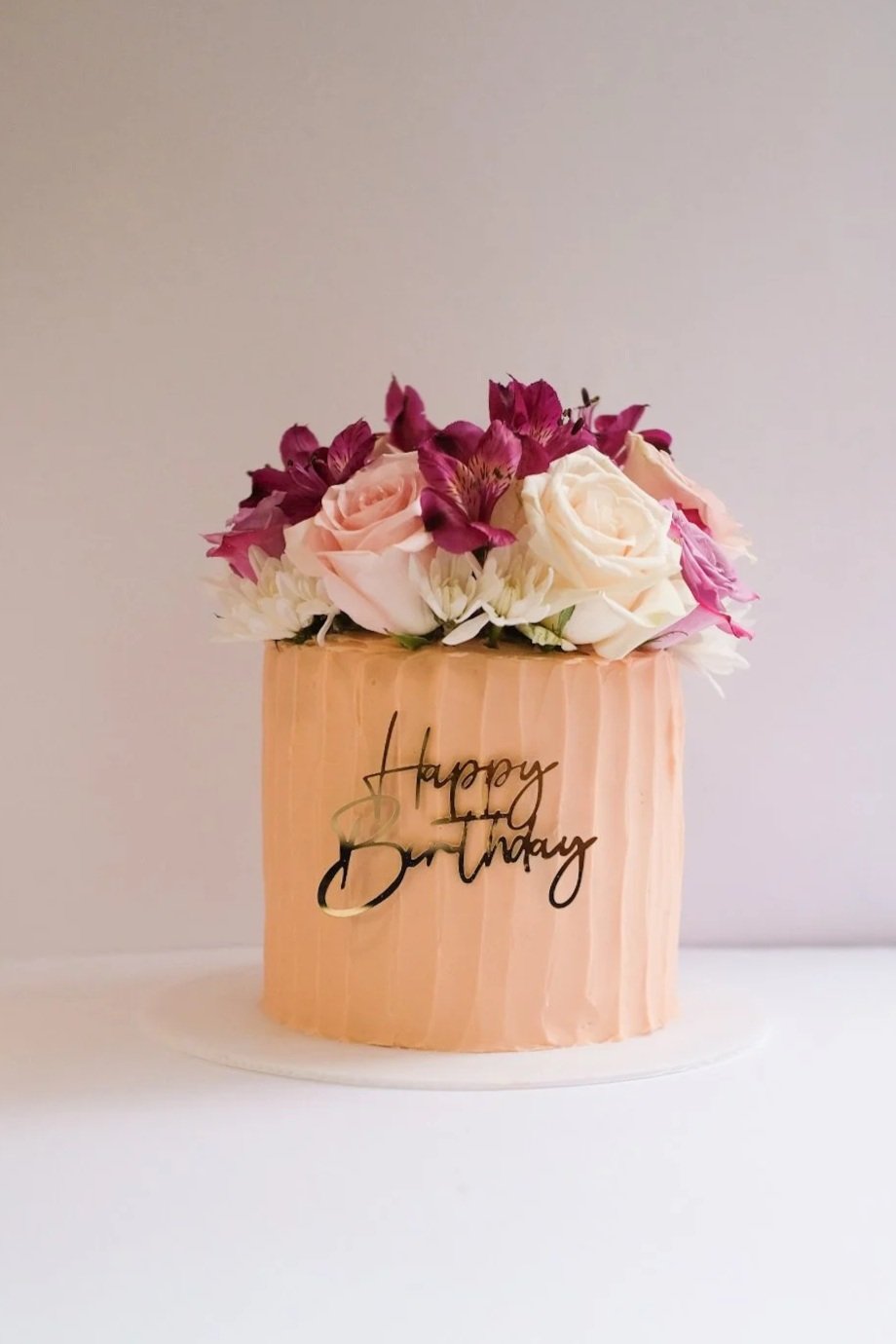 A cake flower arrangement with pink and white roses and purple flowers in a pink box with a 'Happy Birthday' message.