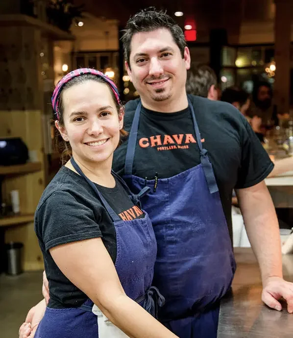 A man and woman standing together in a restaurant kitchen, both wearing black T-shirts with orange text and blue aprons, smiling at the camera. Ilma Lopez for Portland Maine City Council District 5