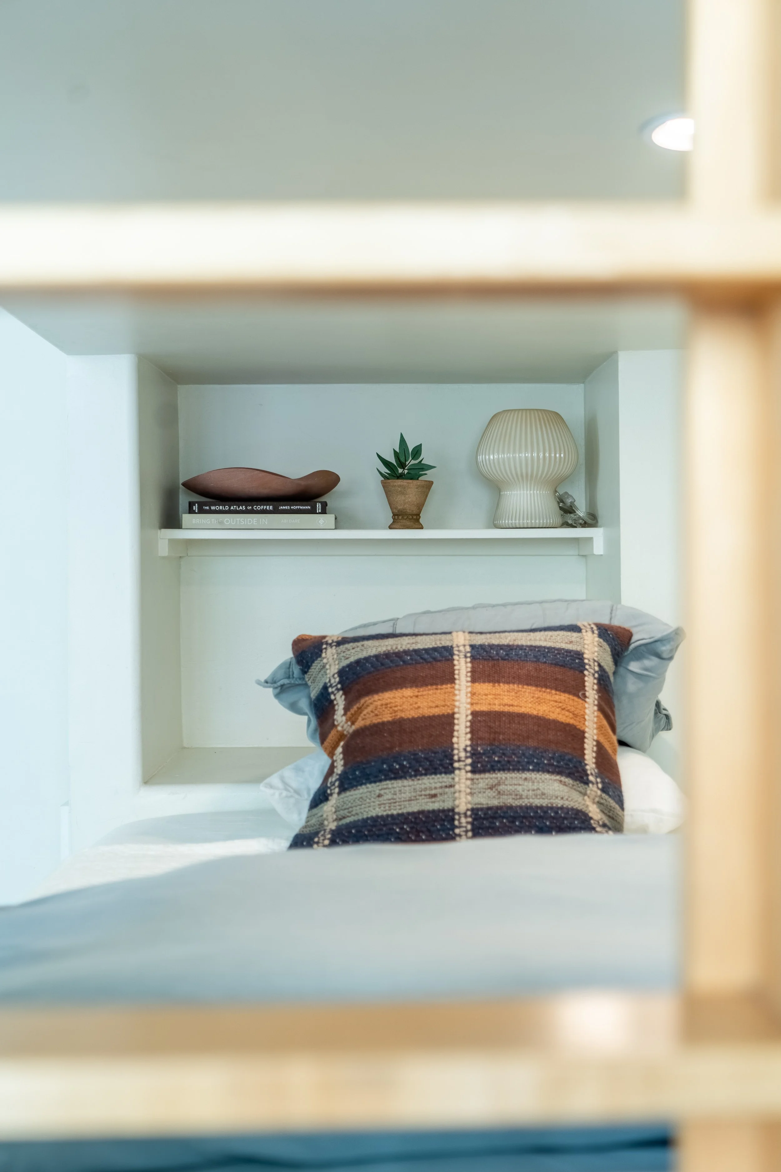 Styled built-in bunk nook at The Solana House with layered bedding, a plaid accent pillow, floating shelf, books, and coastal decor.