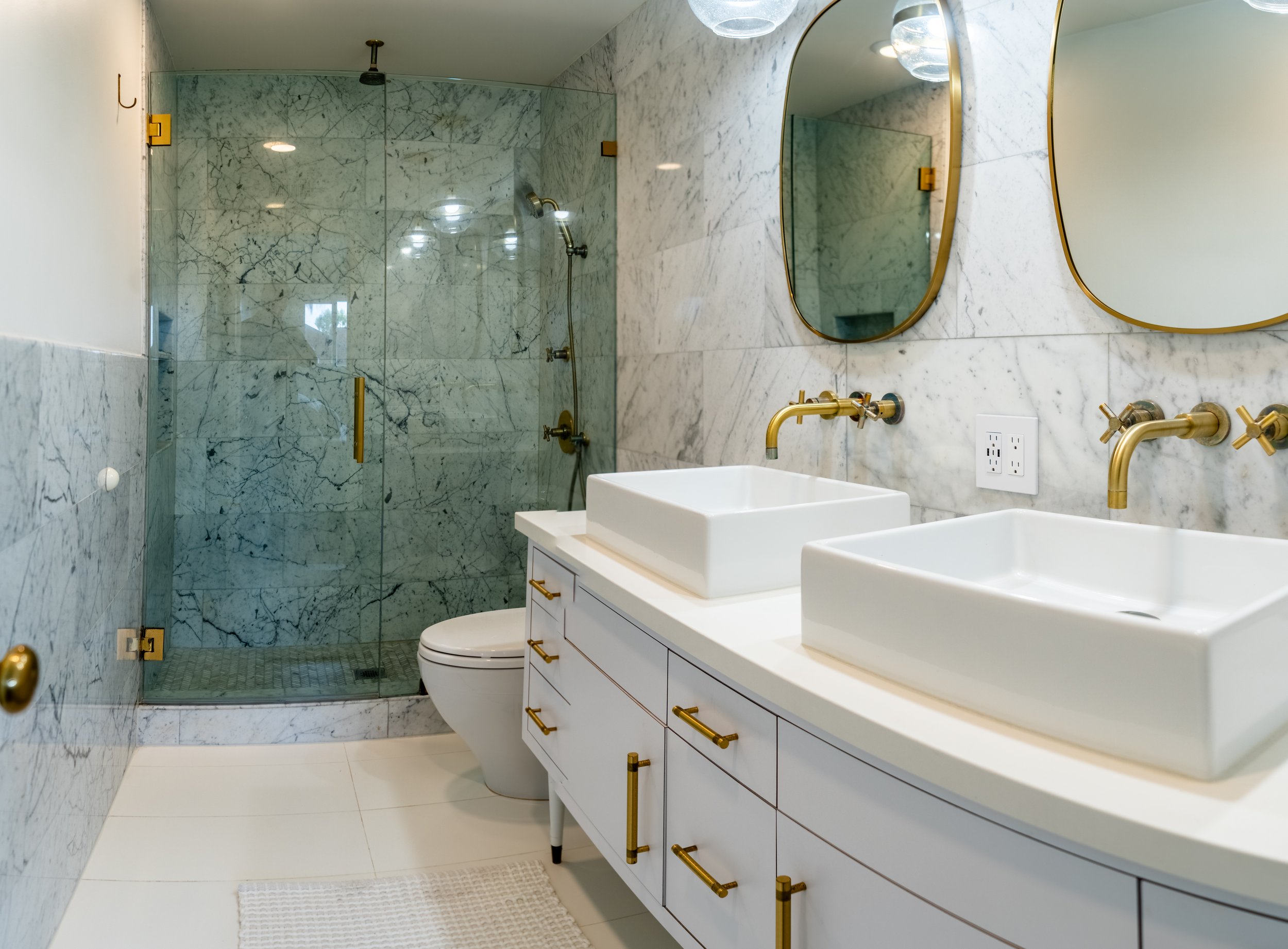 Bright primary bathroom at The Solana house with a double vanity, brass fixtures, oval mirrors, and a glass enclosed marble shower