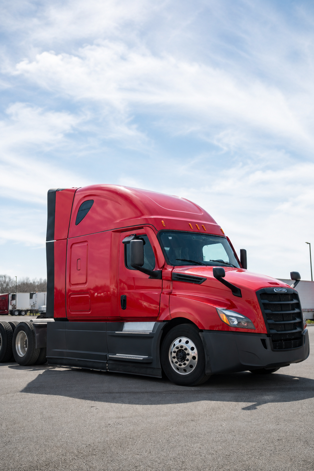 A red semi-truck parked on a paved lot under a partly cloudy sky.