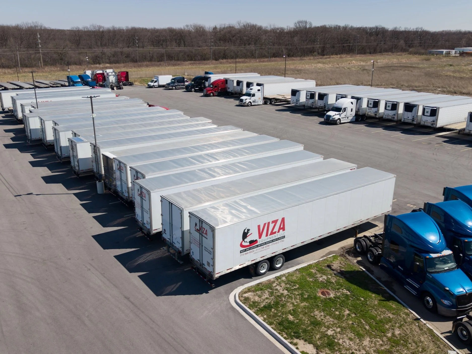 Aerial view of a parking lot with numerous white semi-truck trailers parked in front of a row of trucks and parked cars, with a wooded area in the background.