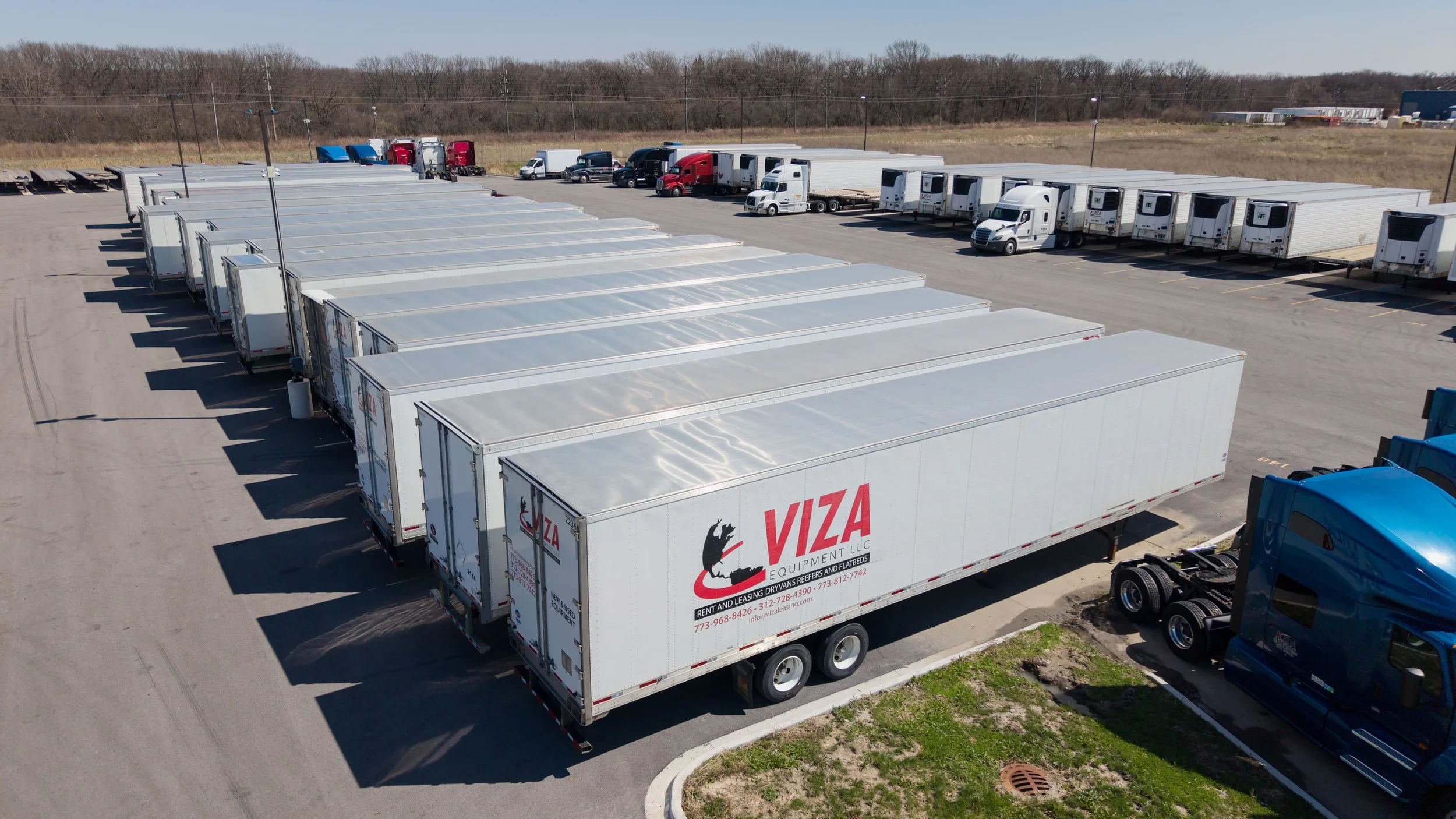 Large parking lot filled with semi-trucks, including white tractor-trailers with VIZA Equipment LLC branding, parked in organized rows on a paved lot surrounded by a grassy area and a line of trees in the background.