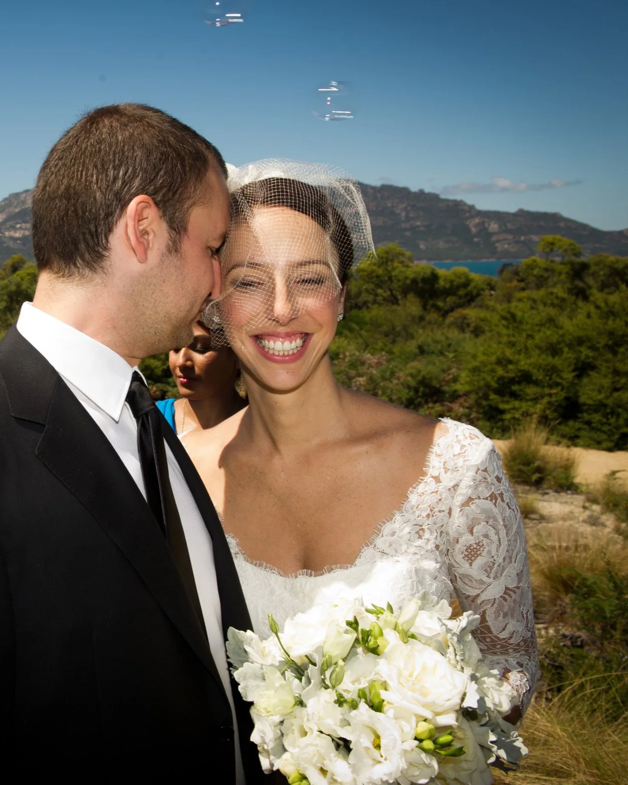 Newlywed couple smiling, with the woman in a lace wedding dress and veil holding a bouquet of white flowers, outdoors with greenery, mountains, and a blue sky in the background.