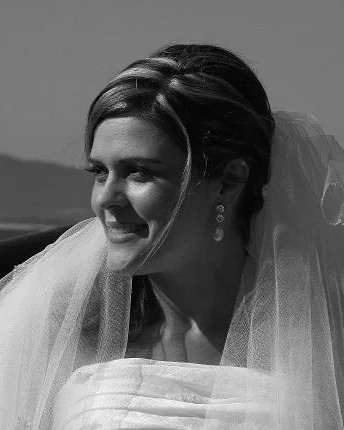 Black and white photo of a smiling bride with styled hair, wearing earrings and a veil, outdoors with a blurred landscape background.