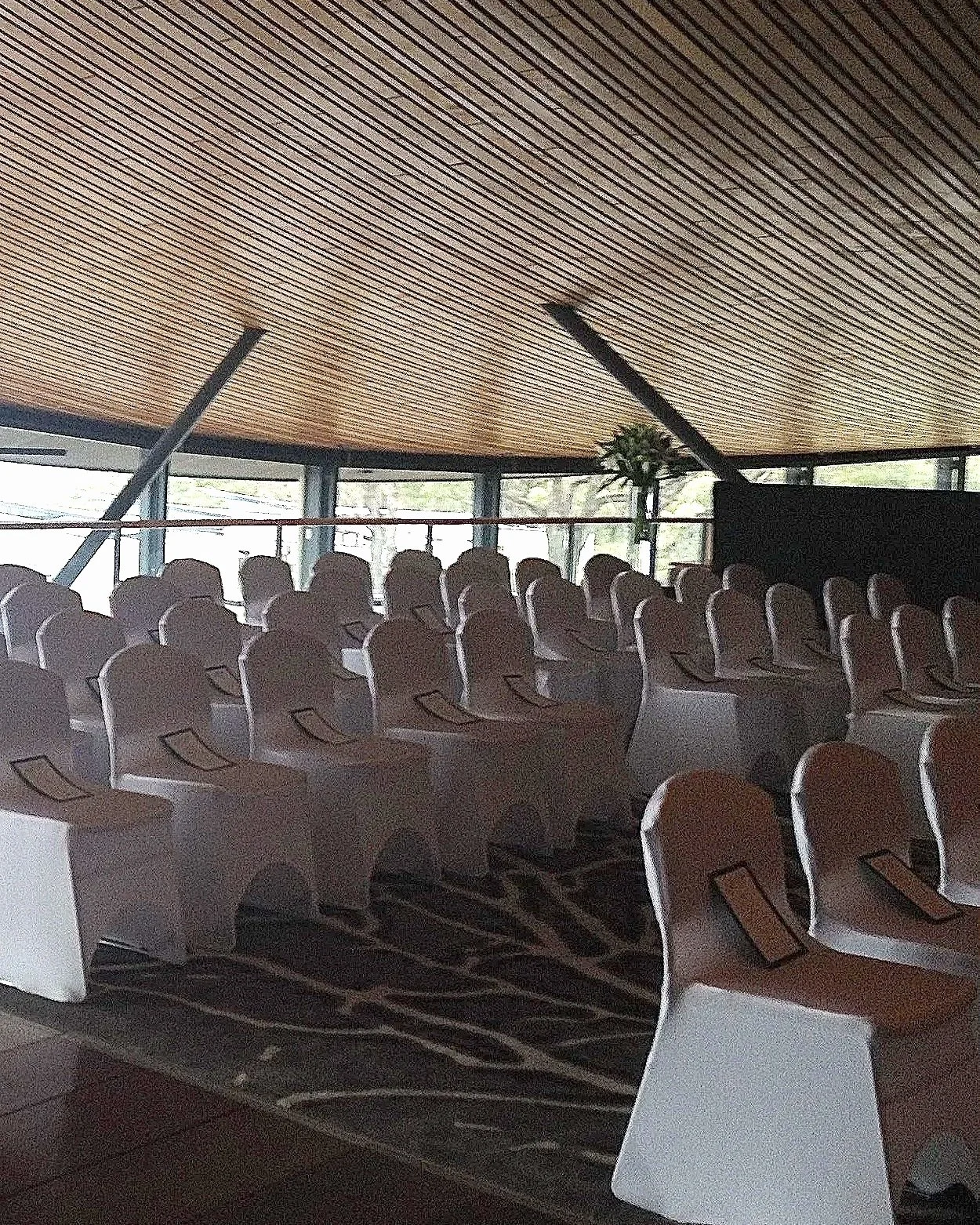Empty conference room with white chairs arranged in rows, a grand piano, and large windows with wood-paneled ceiling.