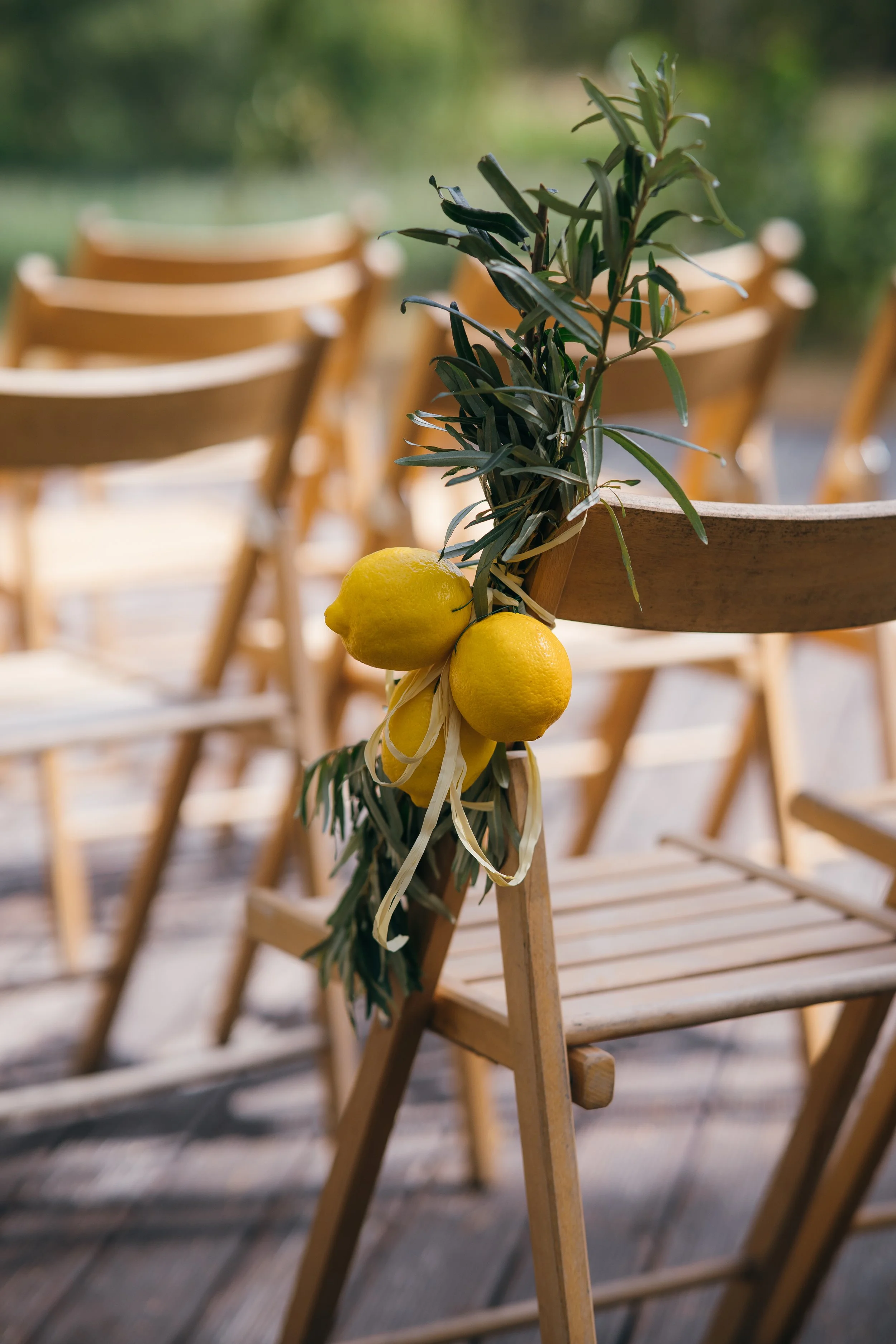 A close-up of a wooden chair decorated with a garland of green leaves and three hanging lemons, with other similar chairs in the background, outdoors.
