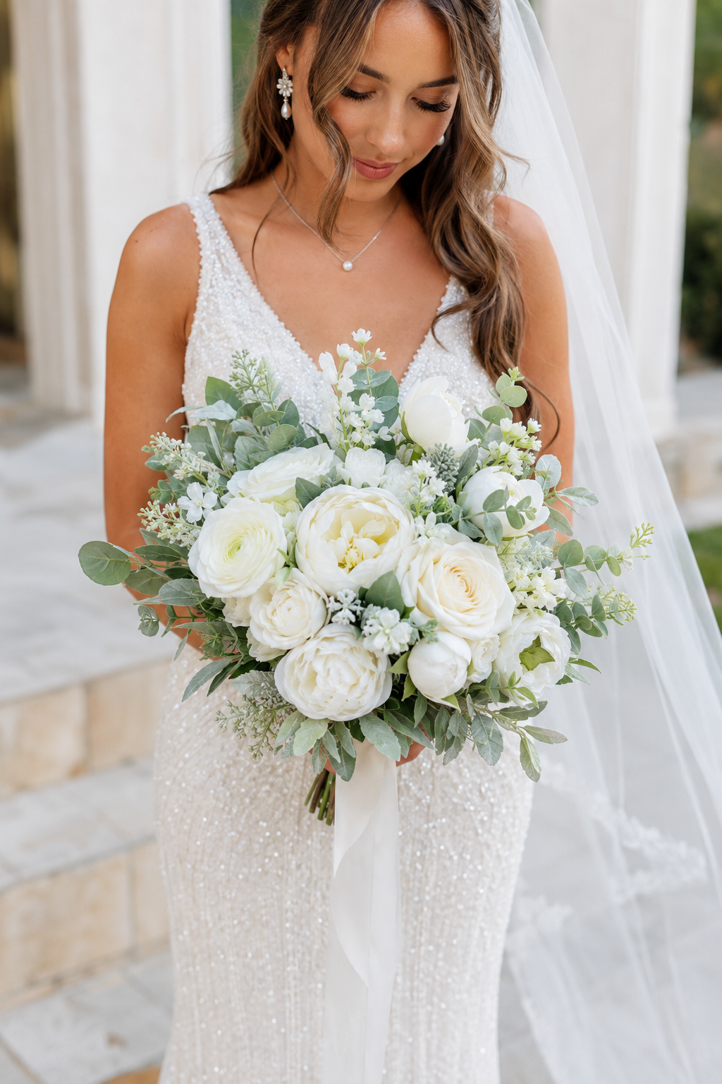 Bride in a beaded white gown holding a large bouquet of white roses, peonies, and greenery, with a veil and outdoor background.