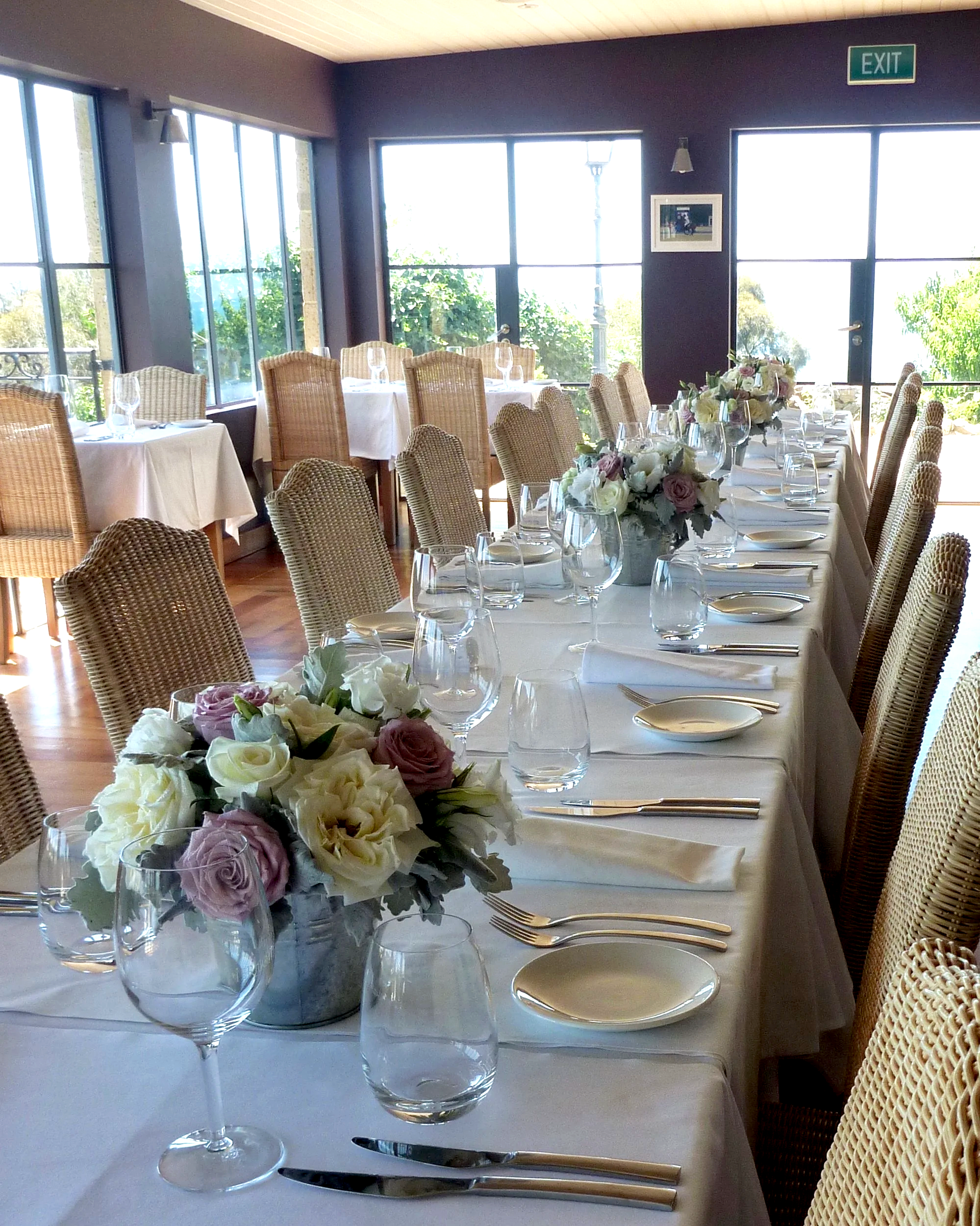Elegant dining room with a long table set with wine glasses, plates, silverware, and floral centerpieces, brightened by large windows showing greenery outside.