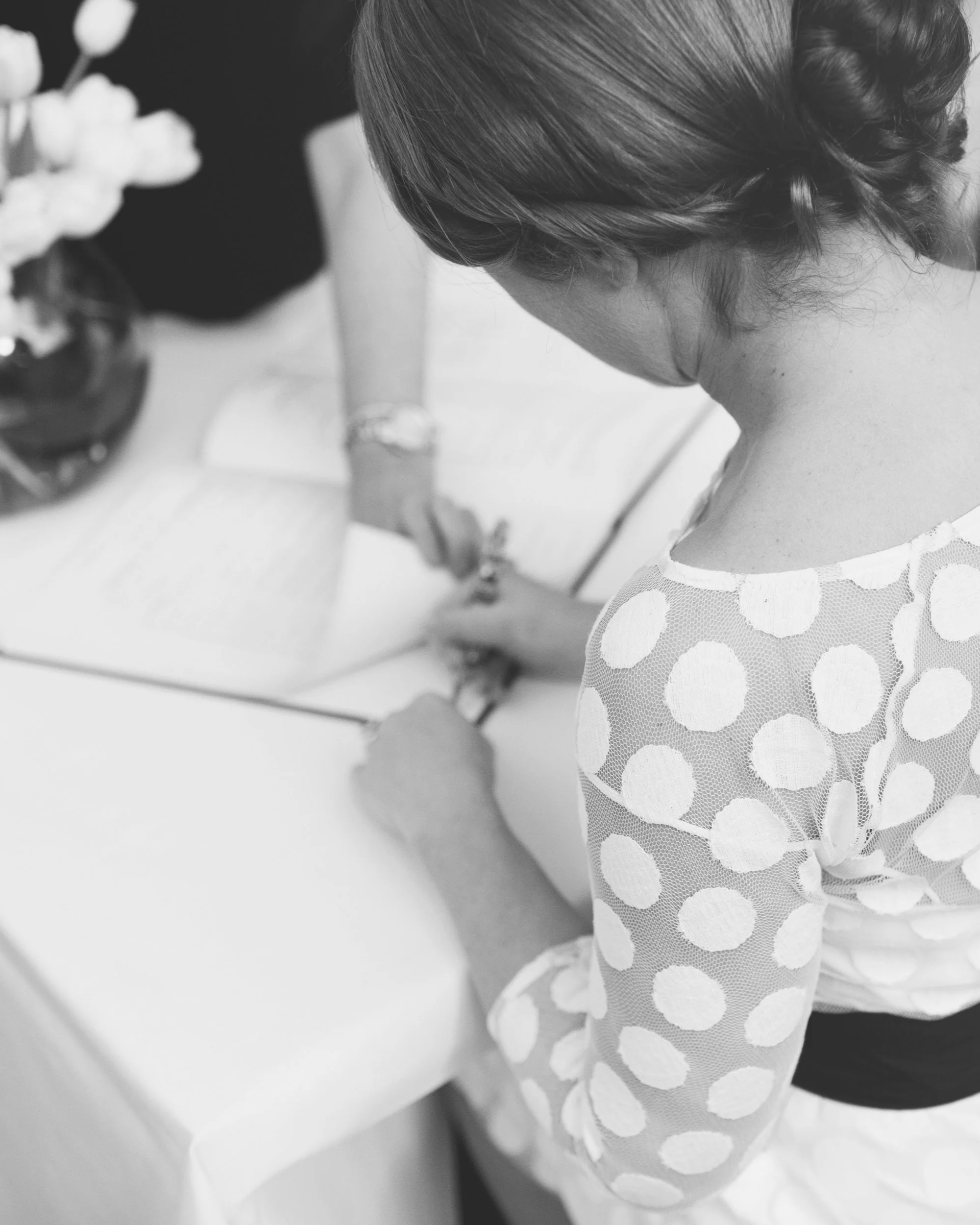A woman with hair styled in an updo, wearing a polka dot dress, standing indoors near a table with a vase of flowers, holding a glass in her hand.