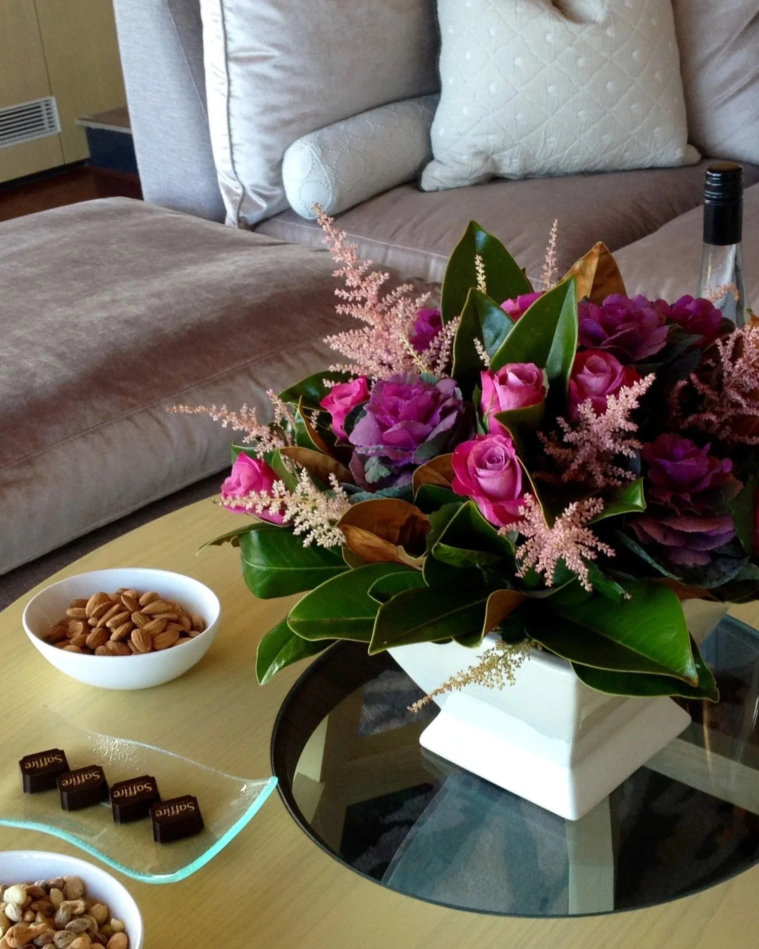 A floral arrangement with pink roses and greenery on a table, a bowl of mixed nuts nearby, and a brown leather couch with cream-colored pillows in the background.