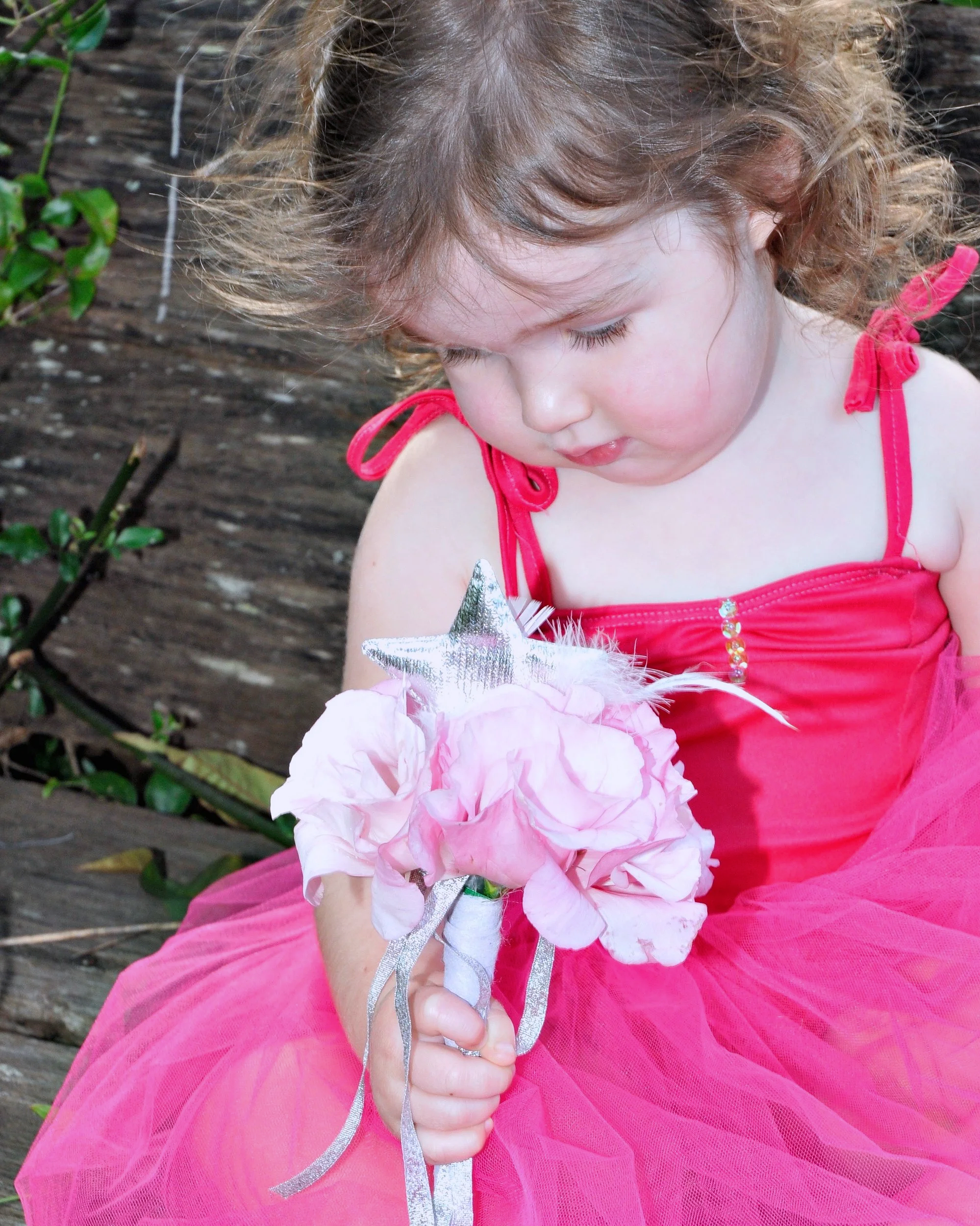 A young girl with curly hair wearing a pink dress holding a bouquet of pink flowers and a silver star-shaped wand, sitting outdoors on a wooden surface.