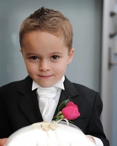 Young boy dressed in a tuxedo with a white bow tie and a pink boutonniere, holding a white object, possibly a cake, during a formal event.