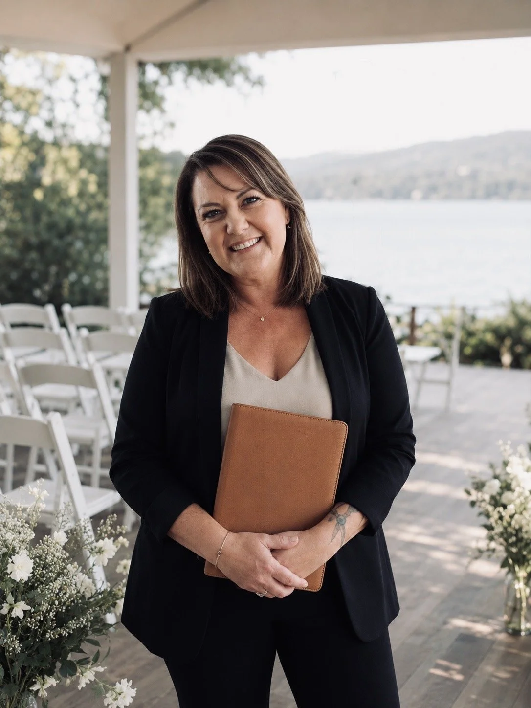 A woman in a black blazer holding a tan notebook, smiling outdoors near a lake with chairs and white flowers around, under a canopy.
