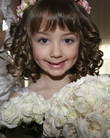 A young girl with curly hair wearing a floral crown and holding a bouquet of white roses, smiling at the camera.