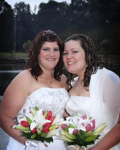 Two women in wedding dresses holding bouquets of pink and white lilies, smiling outdoors near a body of water.
