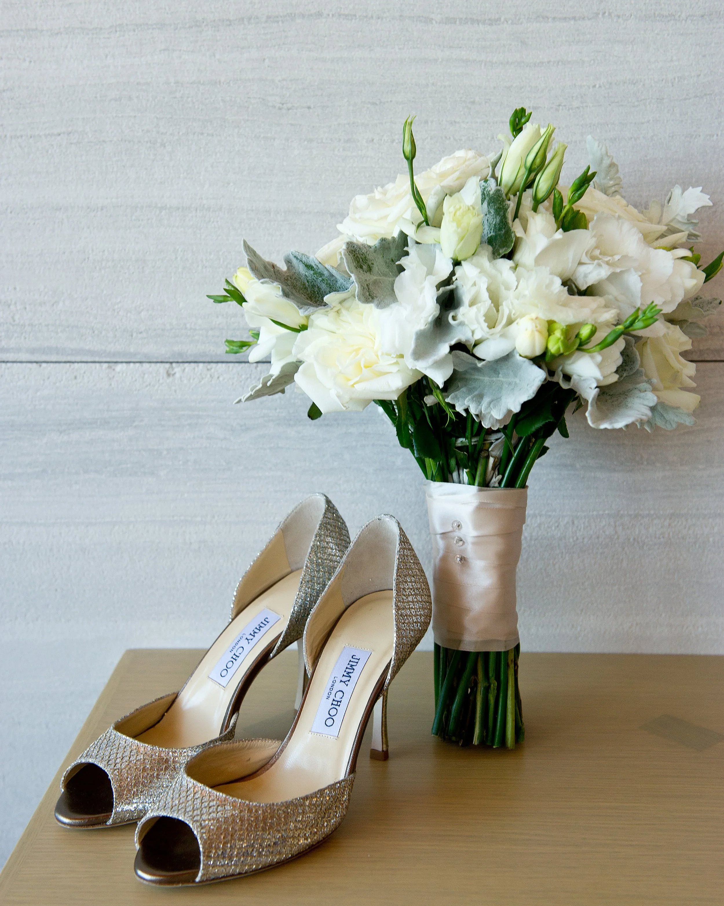 A bouquet of white flowers and green foliage in a wrapped vase, alongside a pair of gold glittery high-heeled shoes on a wooden surface against a light-colored wall.