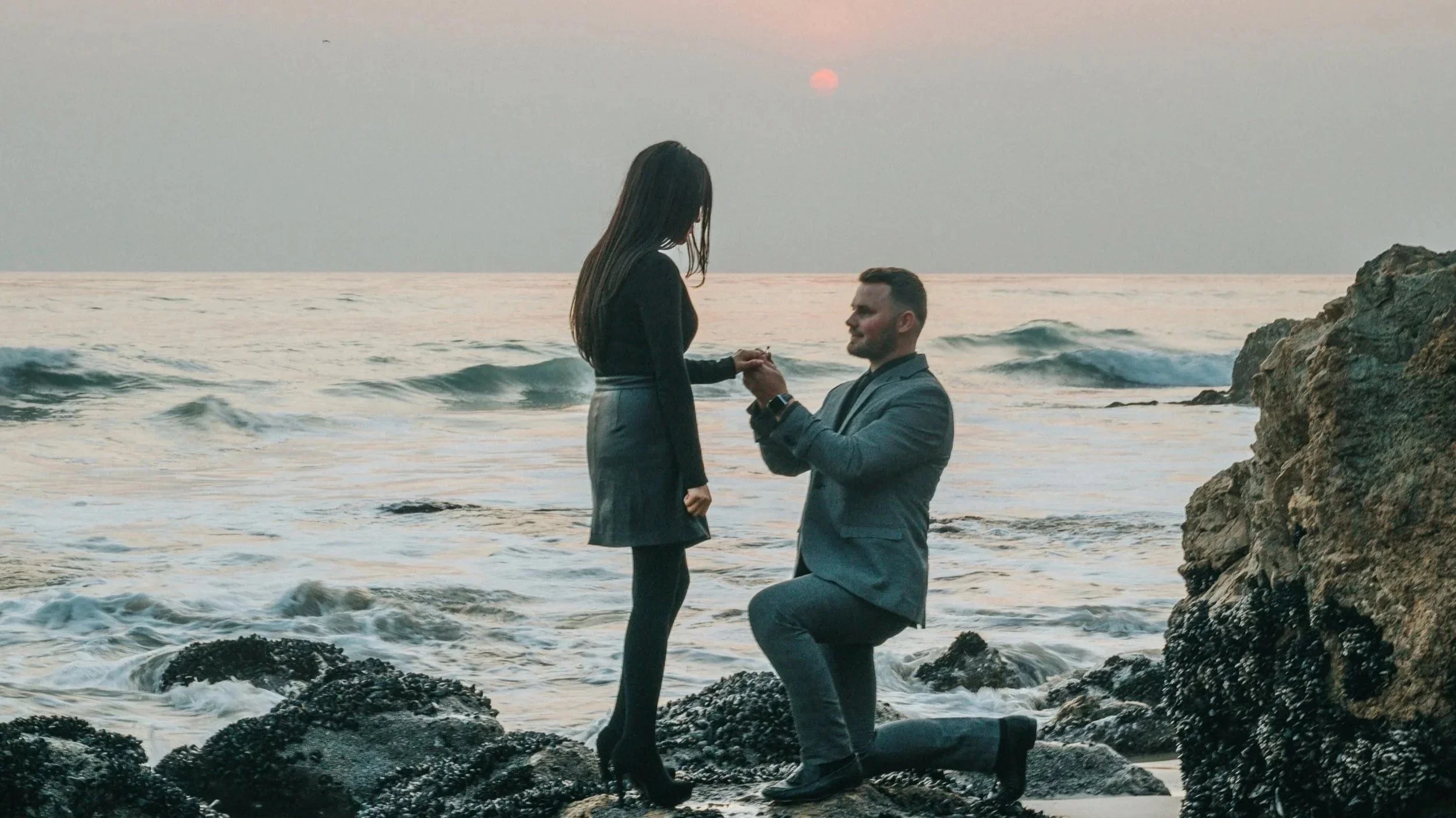 A man in a gray suit proposing to a woman in a black dress on a rocky beach at sunset.