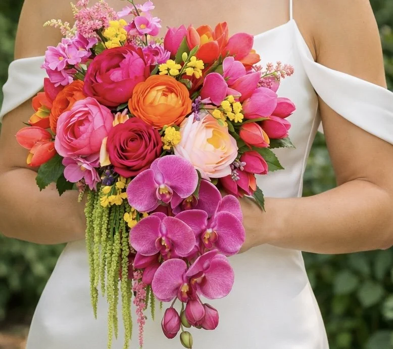 Person holding a vibrant bouquet of pink, orange, yellow, and purple flowers including roses, peonies, tulips, and orchids, dressed in a white off-the-shoulder dress.