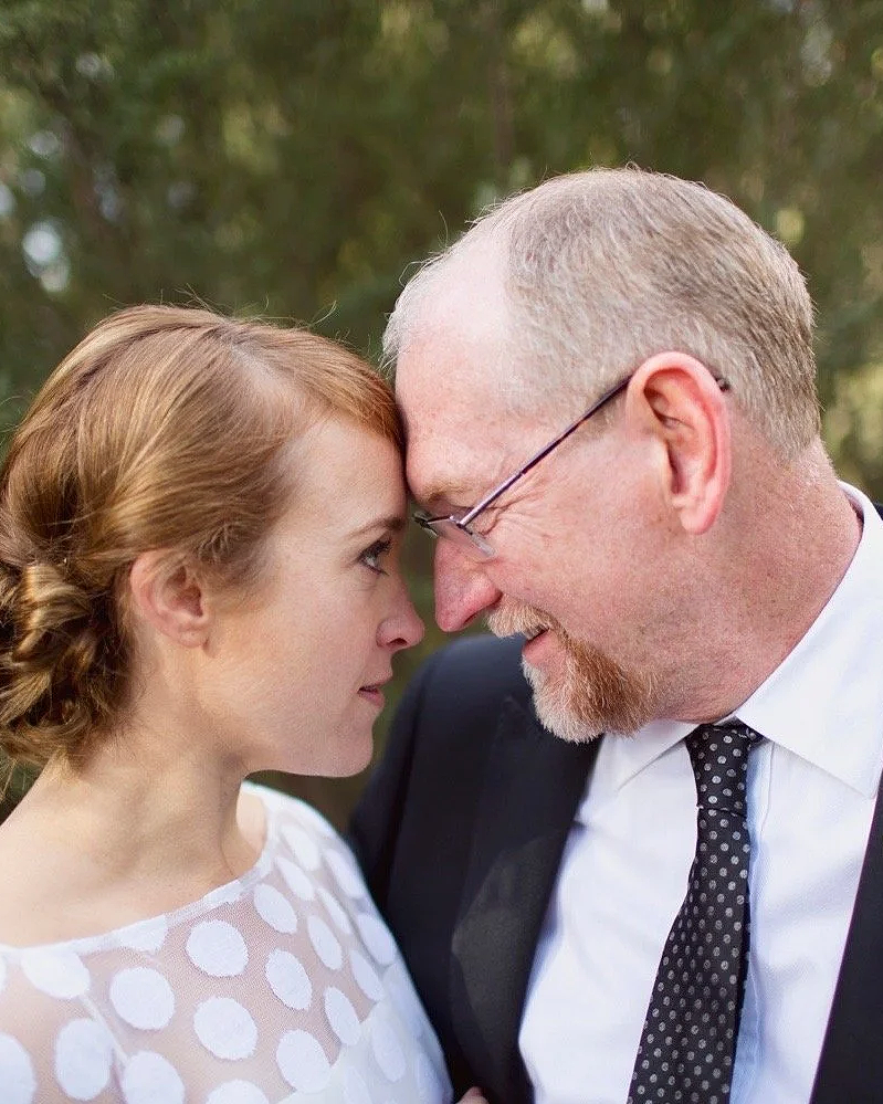 A man and woman face each other closely with foreheads touching, outdoors, with blurred green trees in the background.