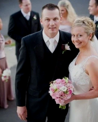 A bride and groom at their wedding, with the bride holding a pink flower bouquet, smiling for the camera.