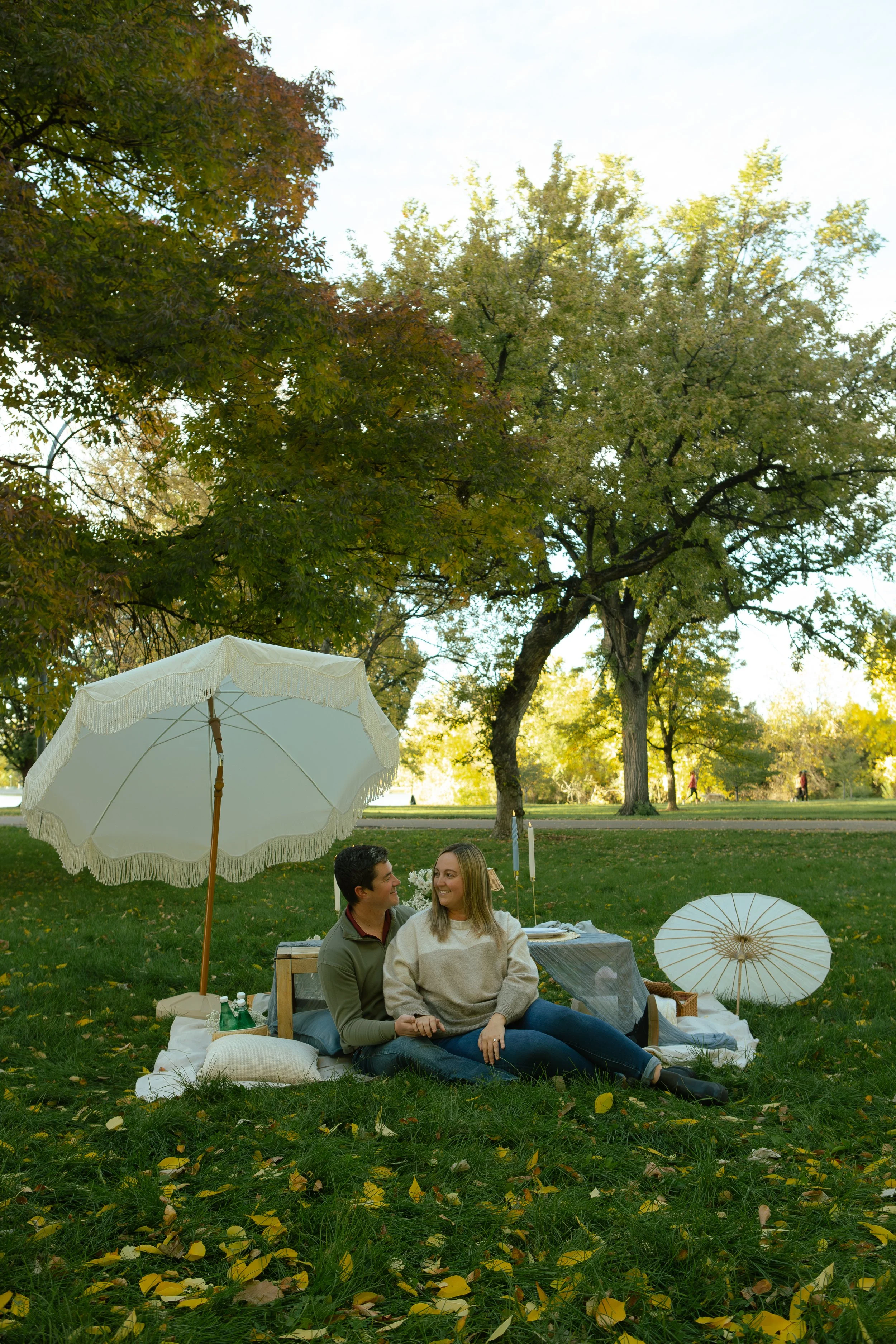 A young couple sitting on a blanket in a park, surrounded by greenery and trees with fall leaves, under white umbrellas, enjoying each other's company.