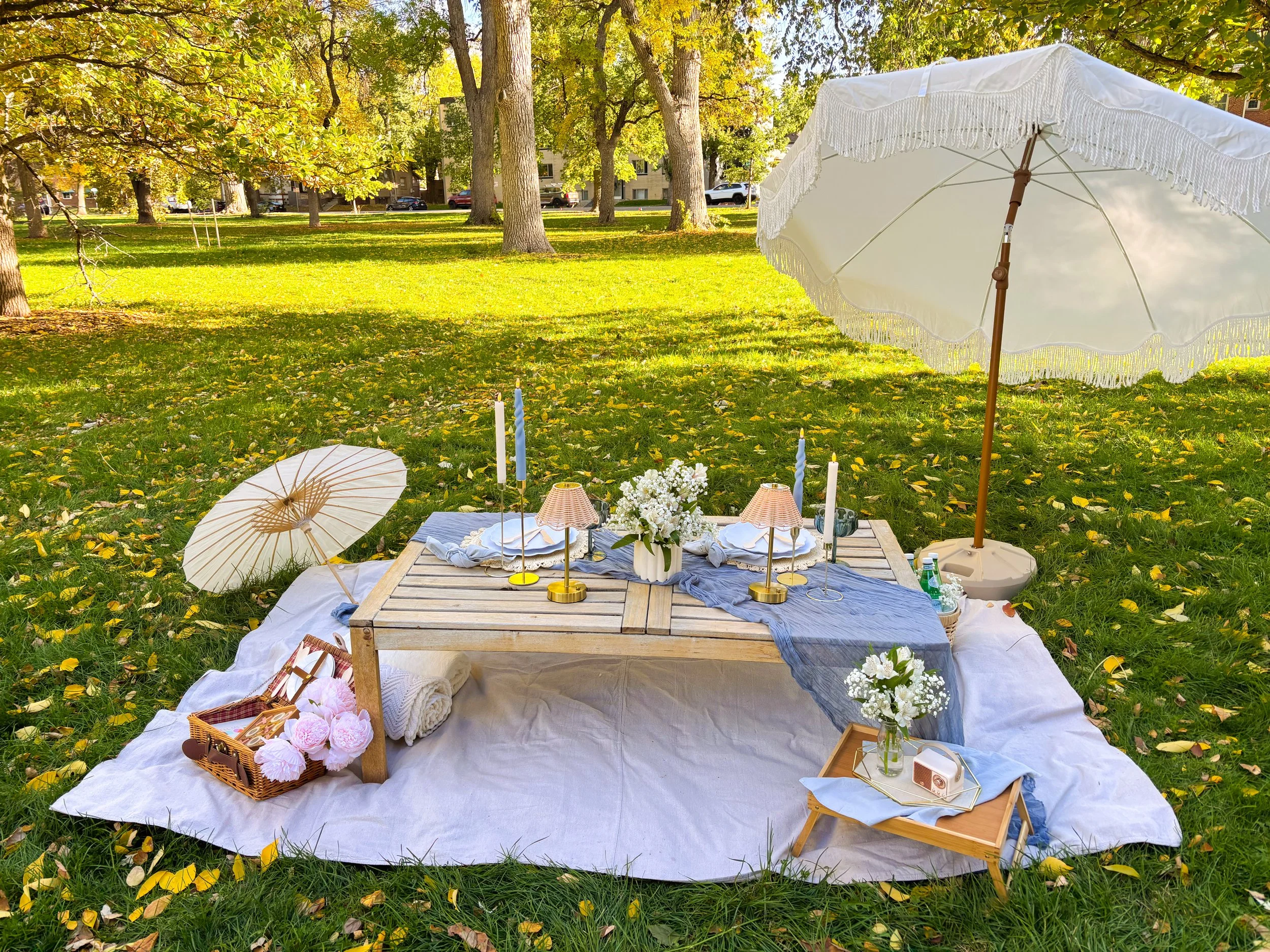 A picnic setup on a white cloth spread on the grass in a park with trees in the background. The setup includes a small wooden table with flowers, candles, and a portable speaker, accompanied by floral arrangements, wicker baskets, umbrellas for decor
