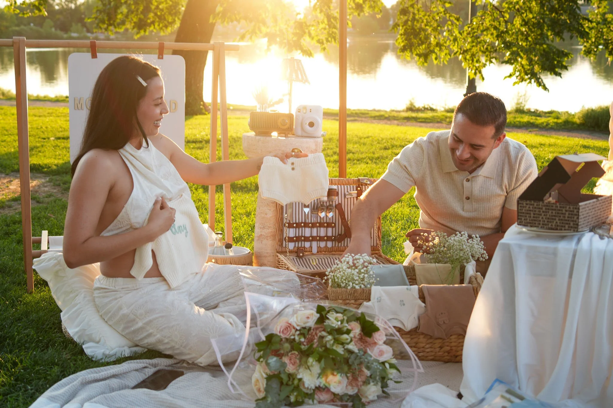 A woman in a white dress sitting on a blanket next to a man at a picnic by a lake during sunset. The woman is smiling and is pregnant. There are flowers, gifts, and cameras on a stand beside them, and trees with green leaves are in the background.