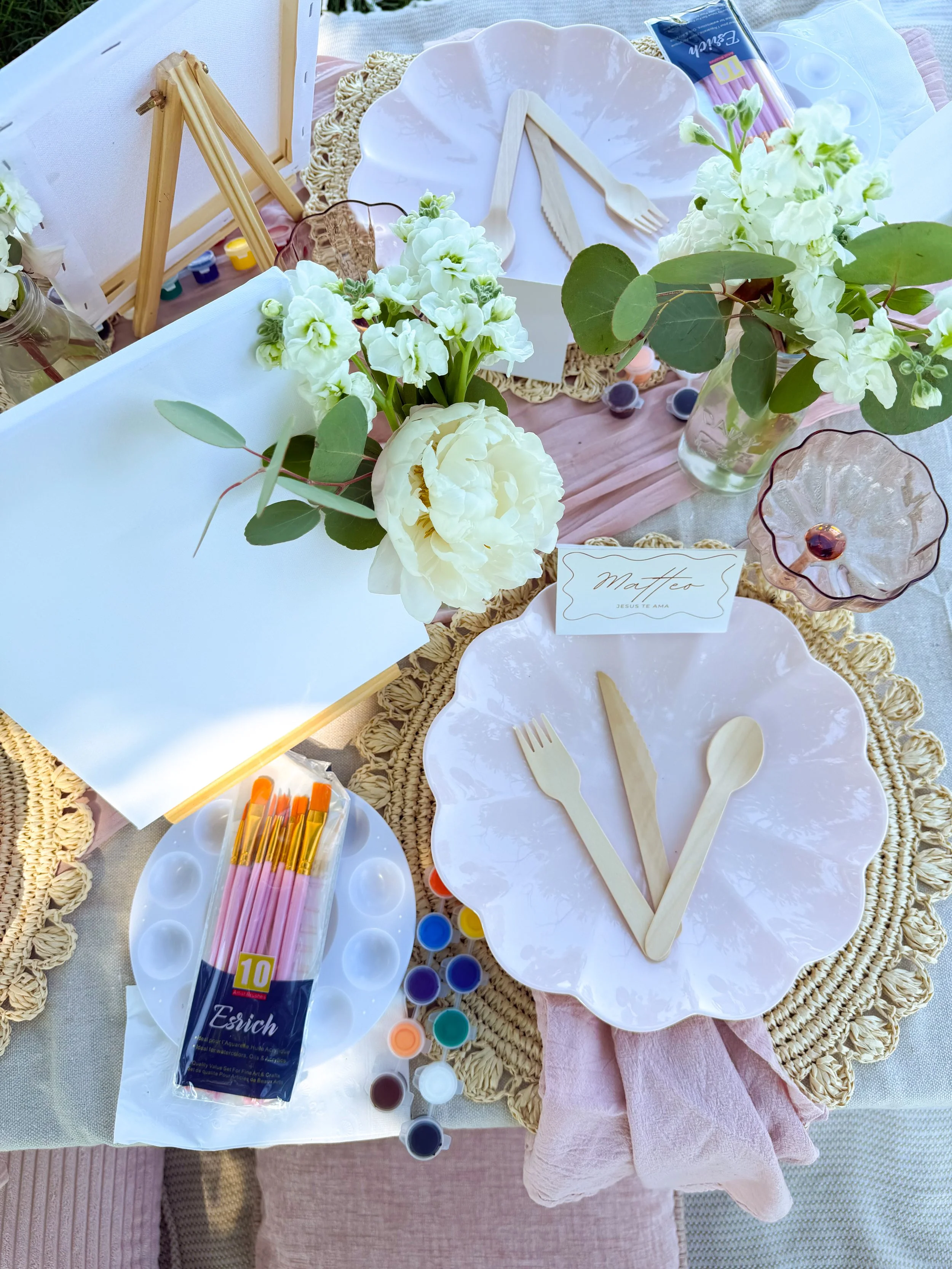 Table decorated with white flowers in glass vases, paper plates, cutlery, and painting supplies, including brushes, on a woven placemat and pink cloth napkin.