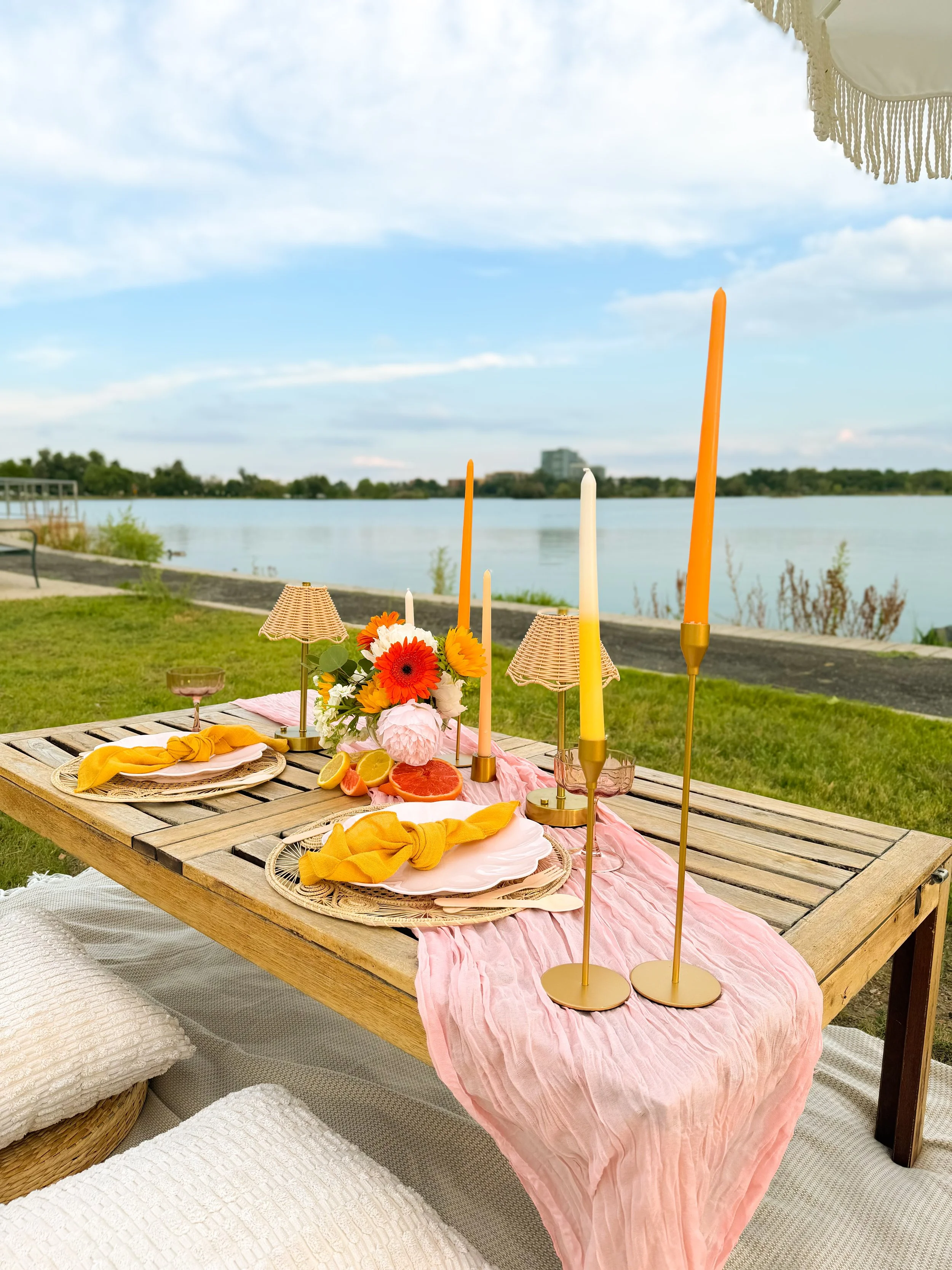A lakeside outdoor dining setup with a wooden table adorned with colorful flowers, tall candles, yellow tableware, set against a backdrop of water, green trees, and a partly cloudy sky.