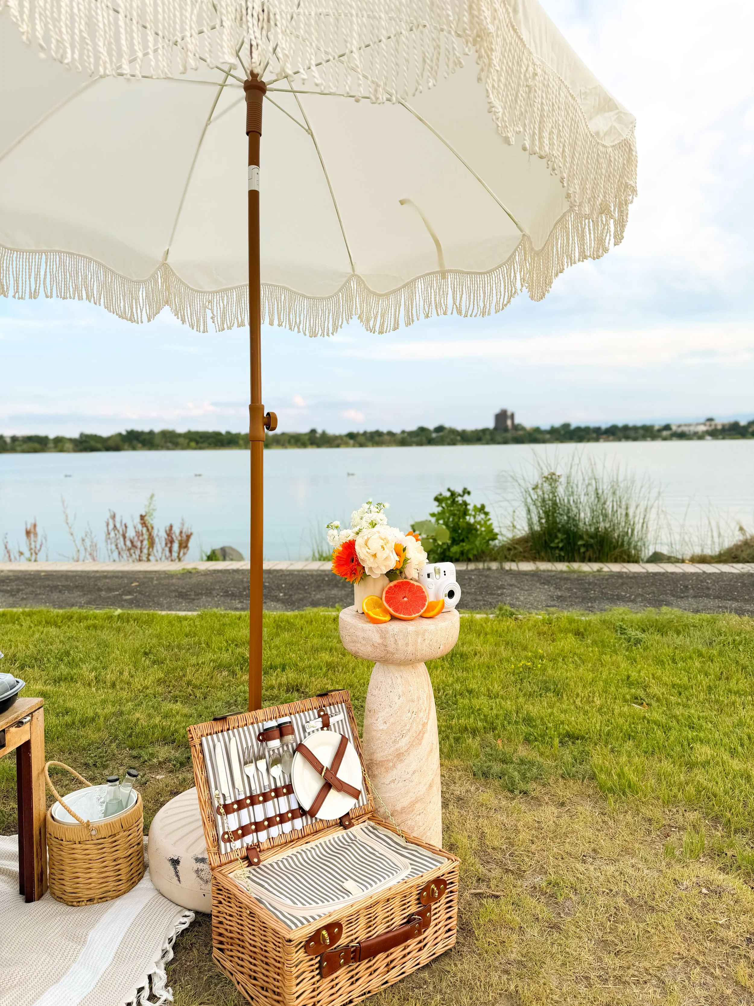 Beachside picnic setup with a large white umbrella, a wicker picnic basket, a basket with drinks, a small round table with a flower arrangement, fruit slices, a vintage camera, and a picnic blanket near the water, with trees and a distant building in