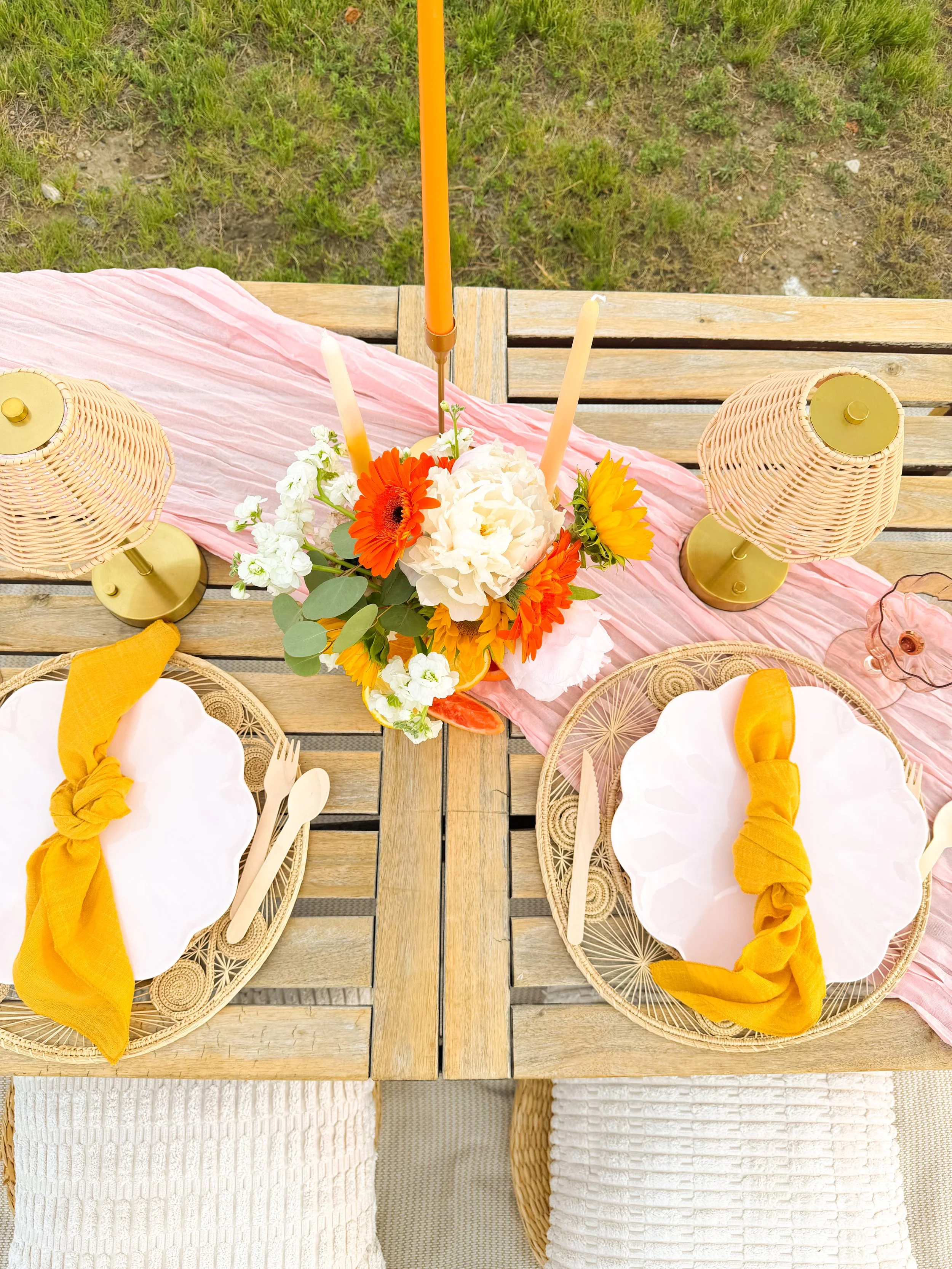 A top-down view of an outdoor dining table with white plates, yellow napkins, yellow utensils, wicker chairs, a pink table runner, and a colorful flower arrangement in a yellow vase. The table is placed on a blanket with grass in the background.