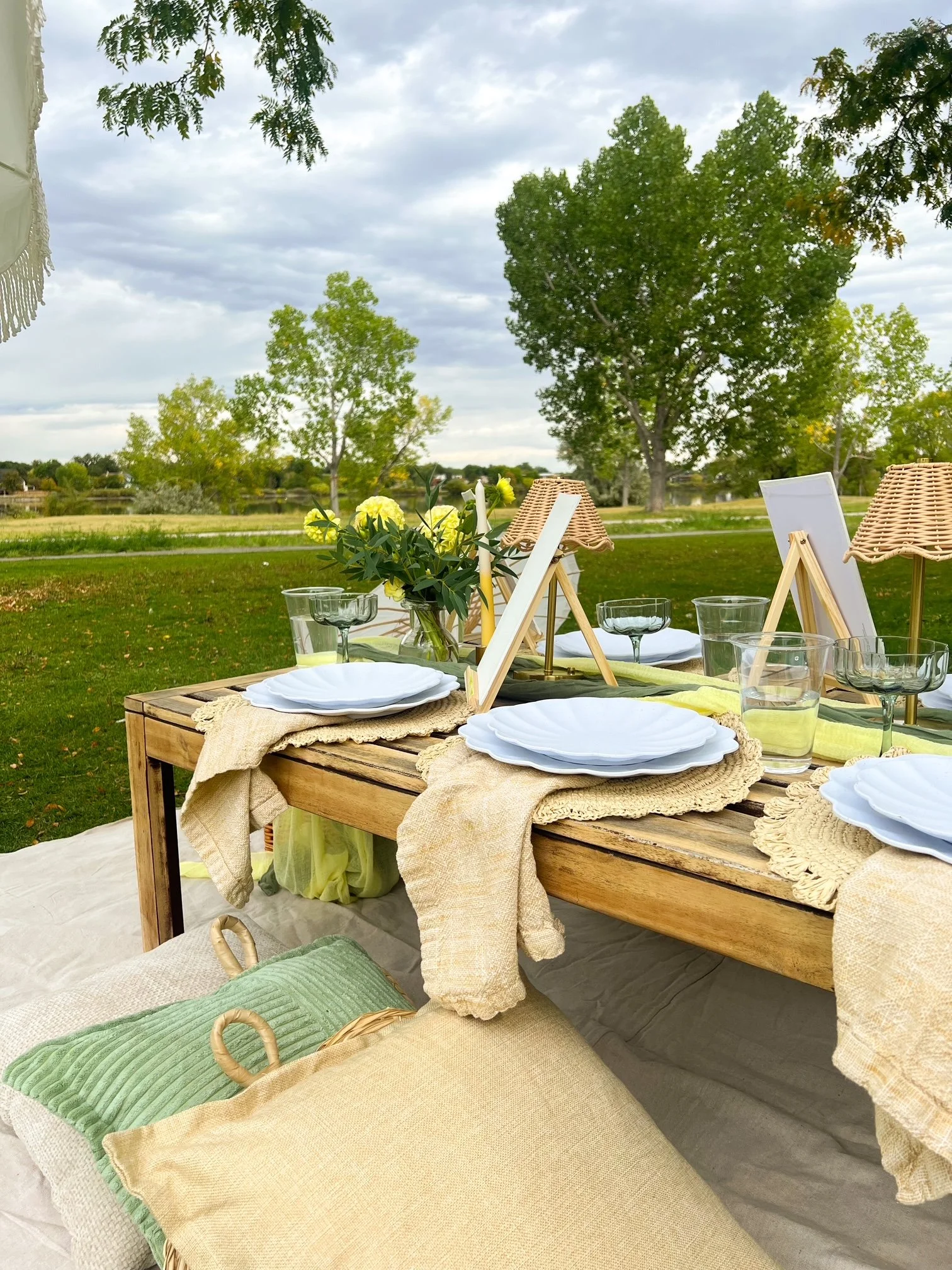 Outdoor dining setup with a wooden table, white plates, glasses, a flower bouquet, and wicker lamps, under trees on a cloudy sky day.