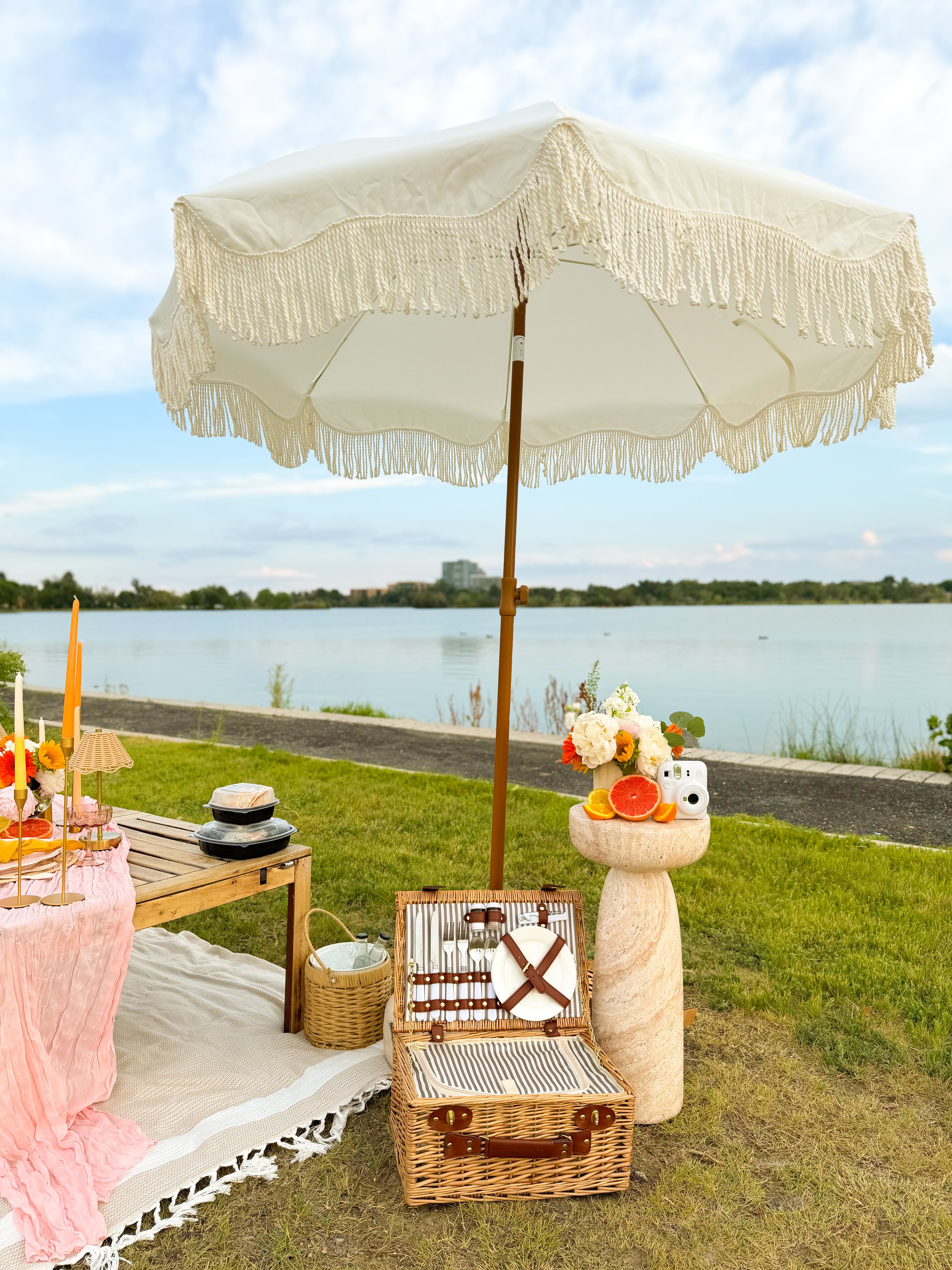 A picnic setup by Sloan's Lake with a white fringed parasol, a table with food and flowers, a wicker picnic basket filled with utensils, and a stone pedestal holding sliced grapefruit and oranges, against a backdrop of water and trees.