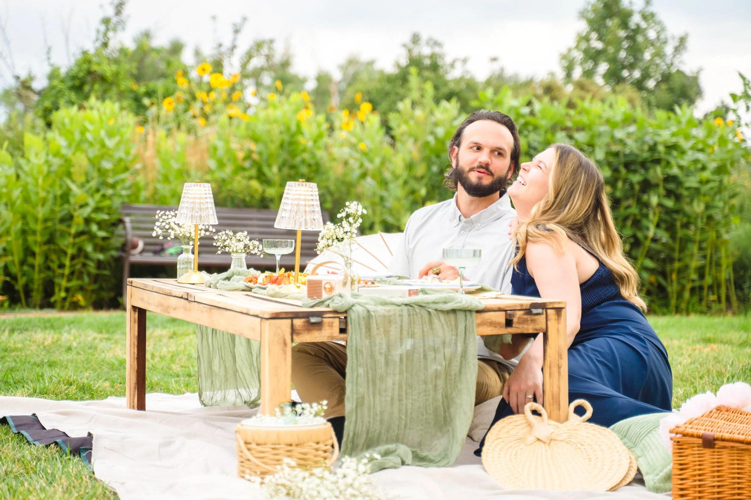 A couple is enjoying a picnic outdoors at a wooden table decorated with flowers, lamps, and tableware, surrounded by lush greenery.