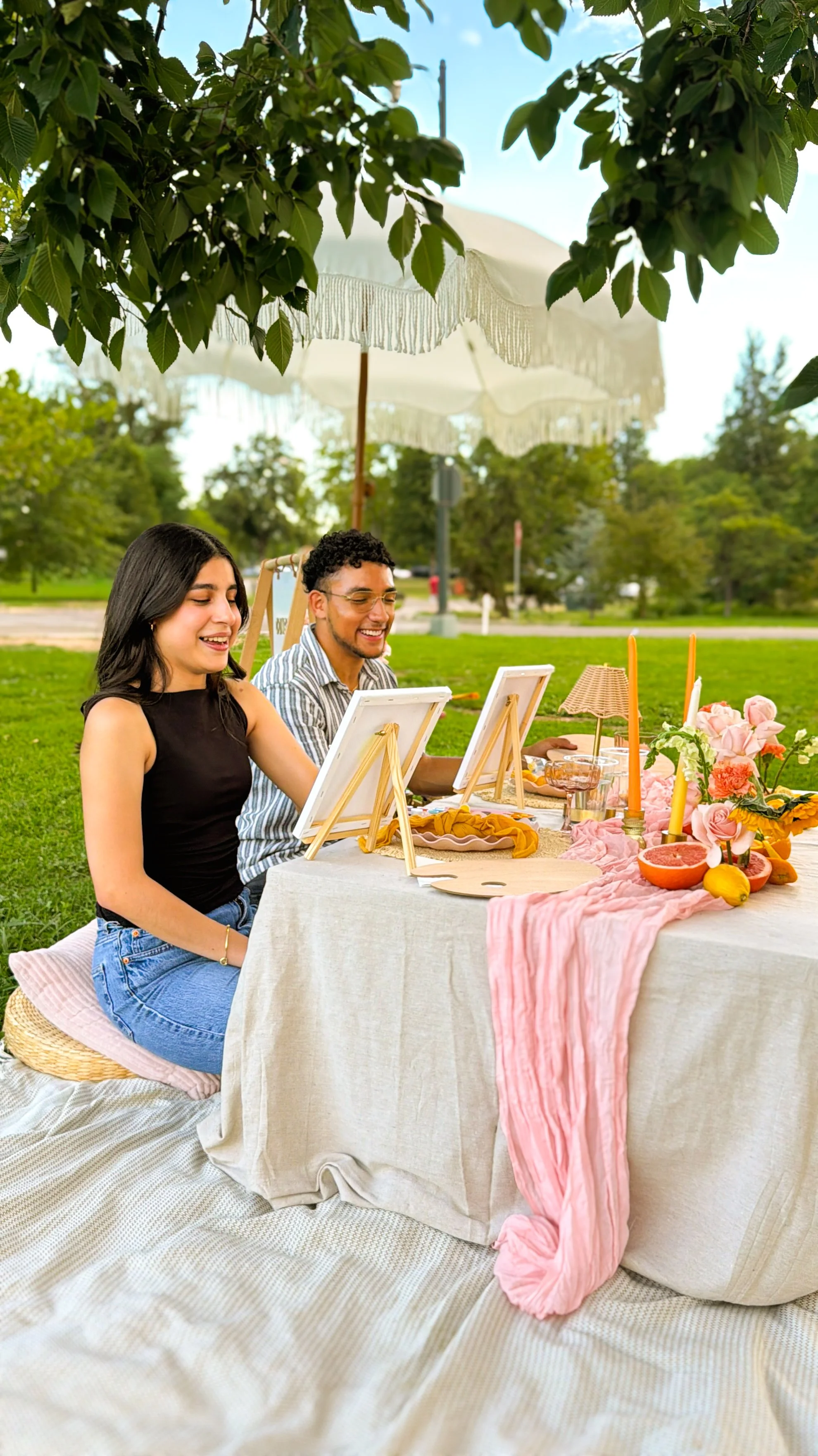 Two people sitting at a table outdoors under a large umbrella, smiling and enjoying a painting activity. The table is decorated with pink flowers, candles, a grapefruit, and a pink cloth. There are trees and a grassy park in the background.