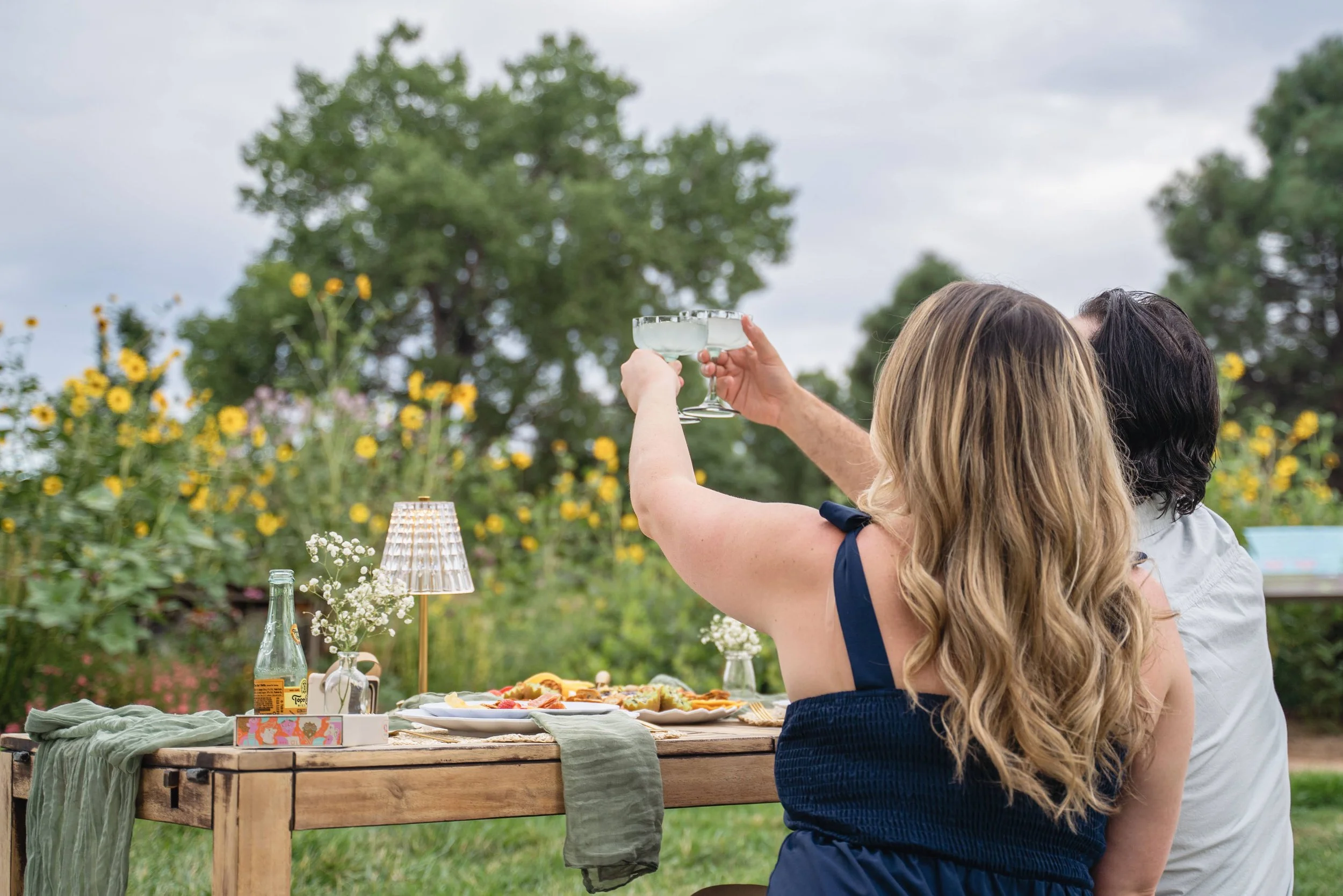 Two people, one with blonde hair and another with dark hair, are toasting with cocktail glasses at a gathering outdoors during daytime. A table with food, drinks, flowers, and decorations is in the foreground, with trees and a cloudy sky in the backg