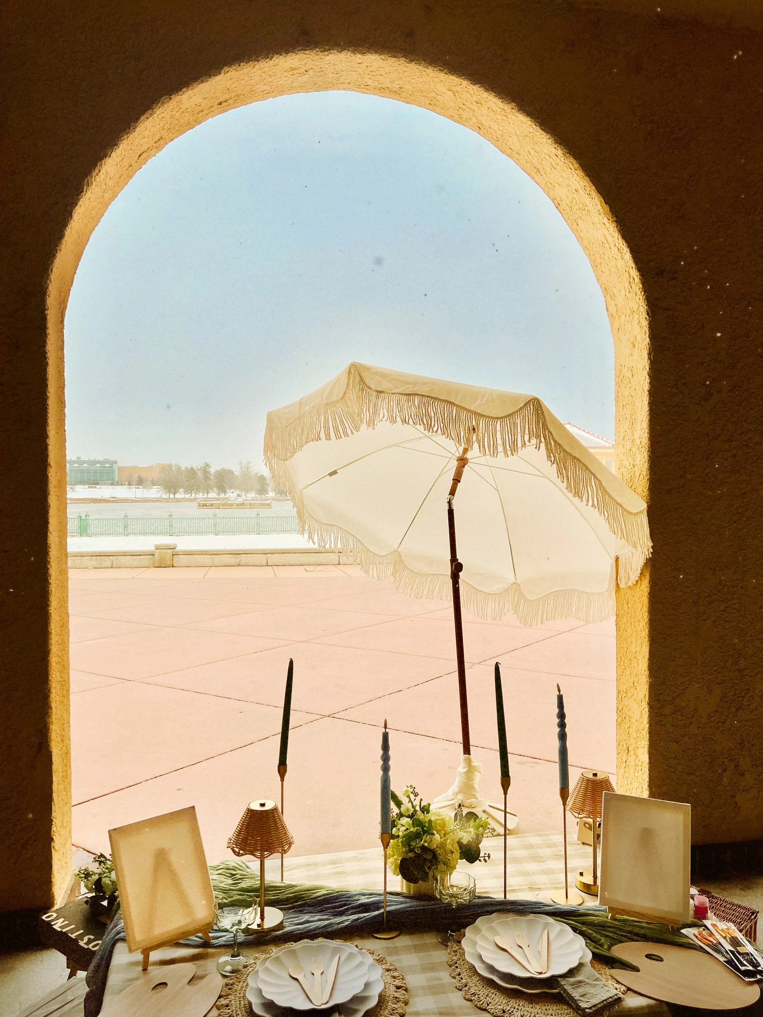 A view from inside a building looking out through a large arch window at a snow-covered outdoor scene with an umbrella and furniture visible in the foreground.
