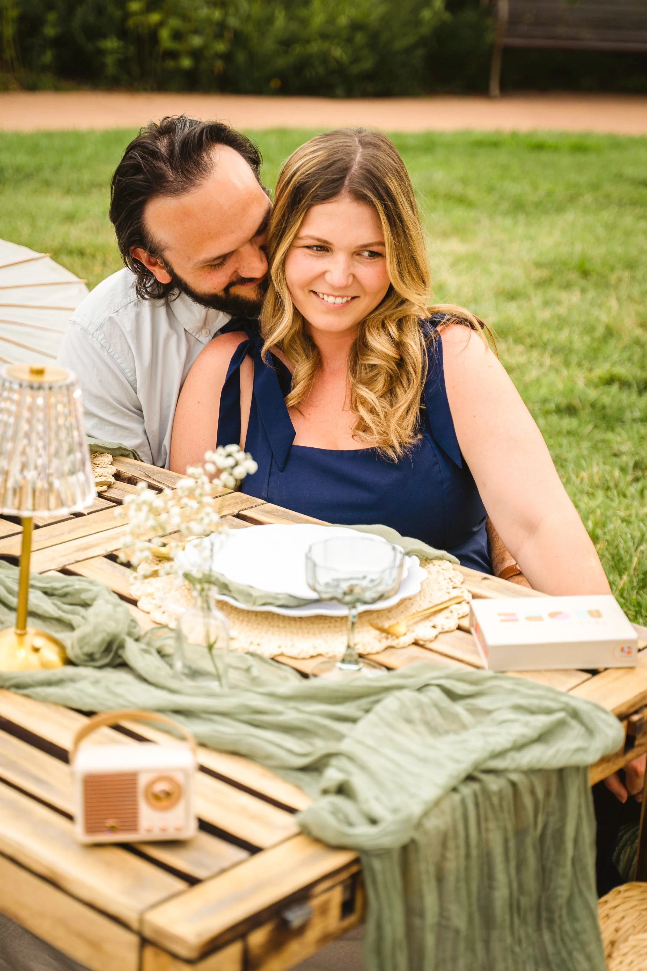 A smiling woman with long wavy blonde hair wearing a navy blue dress, sitting at an outdoor table with a man with dark hair and beard, with his head close to hers. The table has a cloth, flowers, a box, glasses, and a small portable radio. They are o