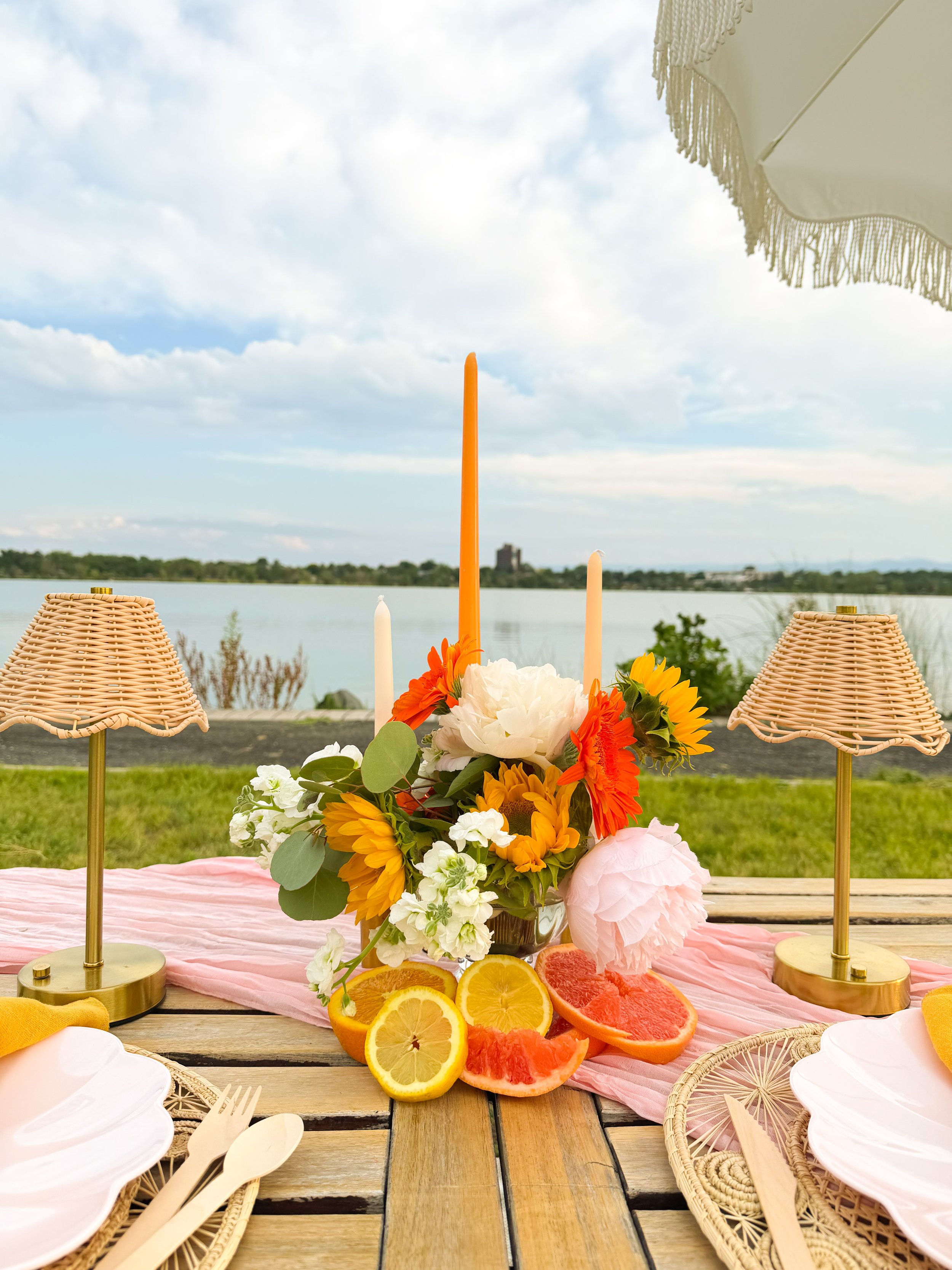 A picnic table set with a pink table runner, wicker lamps, pink and white plates, wooden utensils, a floral centerpiece with orange, white, and yellow flowers and citrus slices, and a view of a lake with greenery and a cloudy sky in the background.