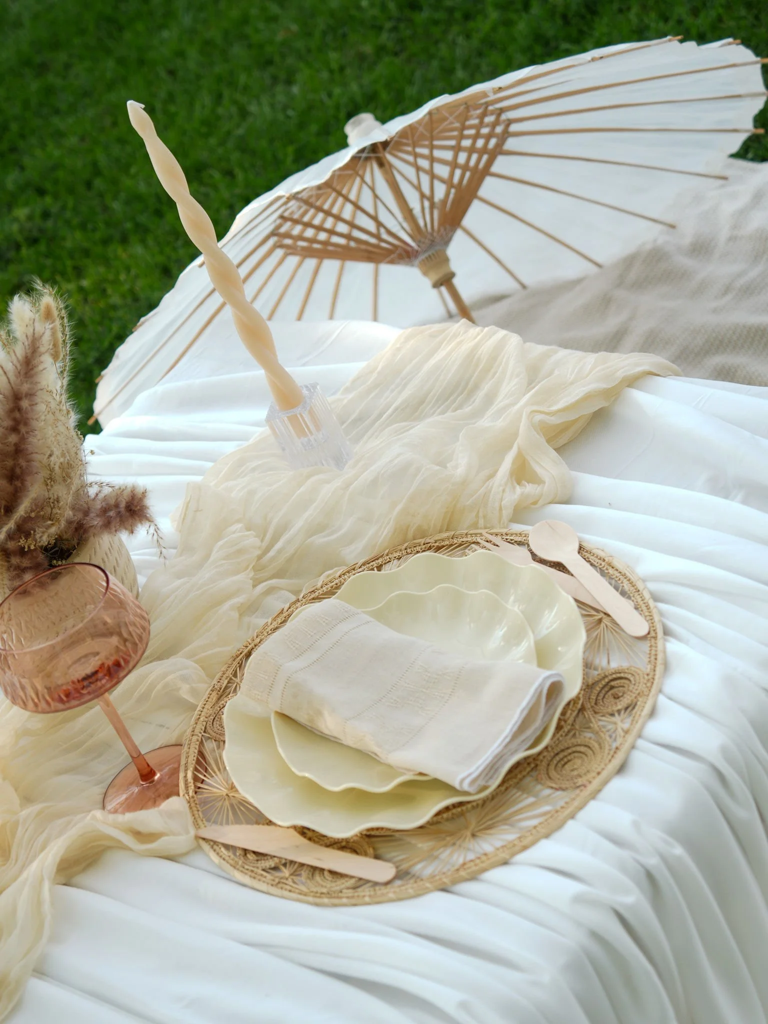 A picnic setup on a white fabric with a wicker tray, stacked white plates, a folded cloth napkin, a pink glass, and a candle. In the background, a white parasol and green grass are visible.